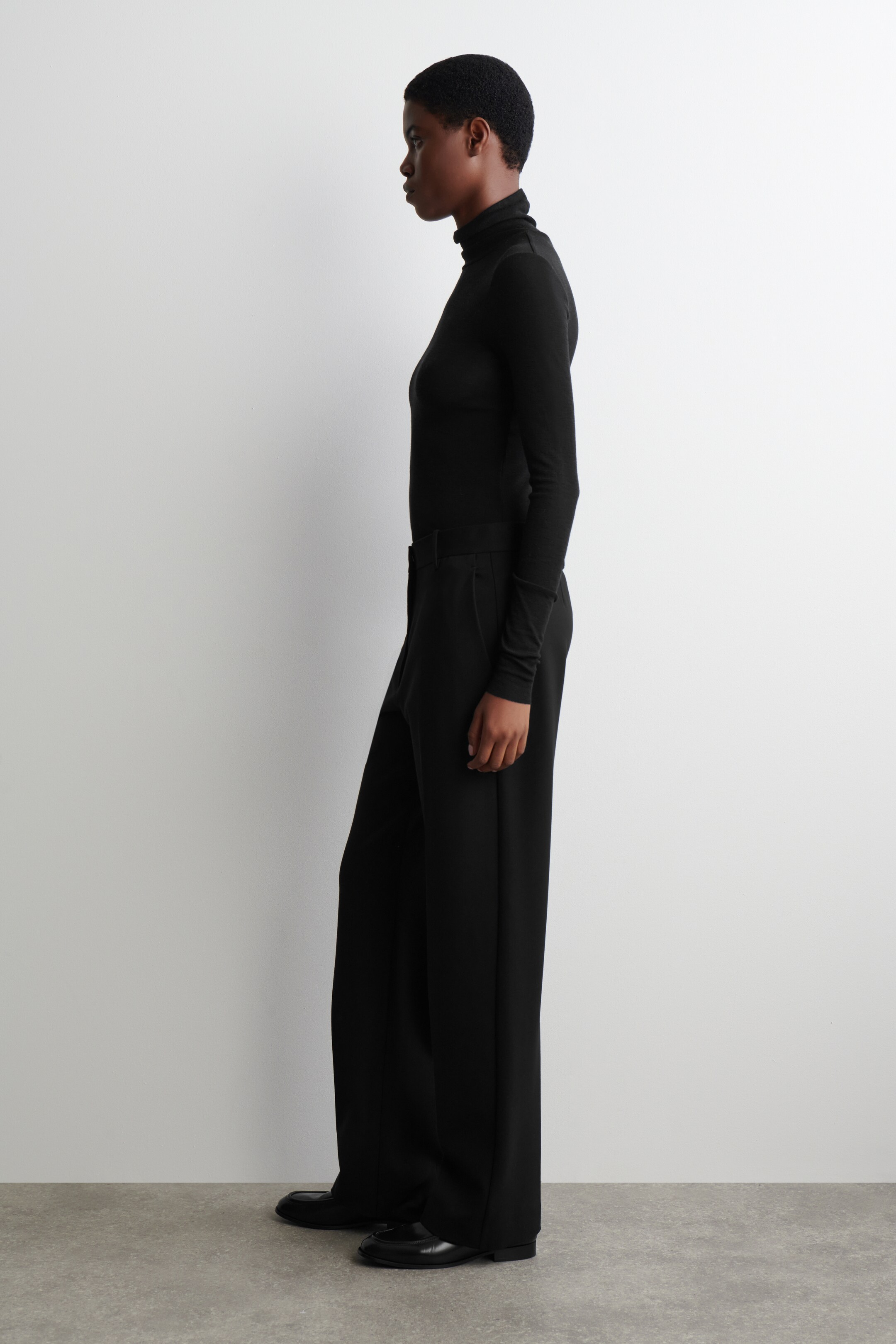 Größeres Bild ansehen: Ein schwarzes, figurbetontes, langärmliges Rollkragenoberteil wird getragen, in schwarze Hosen mit weitem Bein gesteckt. Die Hosen fallen über schwarze flache Schuhe, da die Produkte aus einem Seitenprofil gezeigt werden.