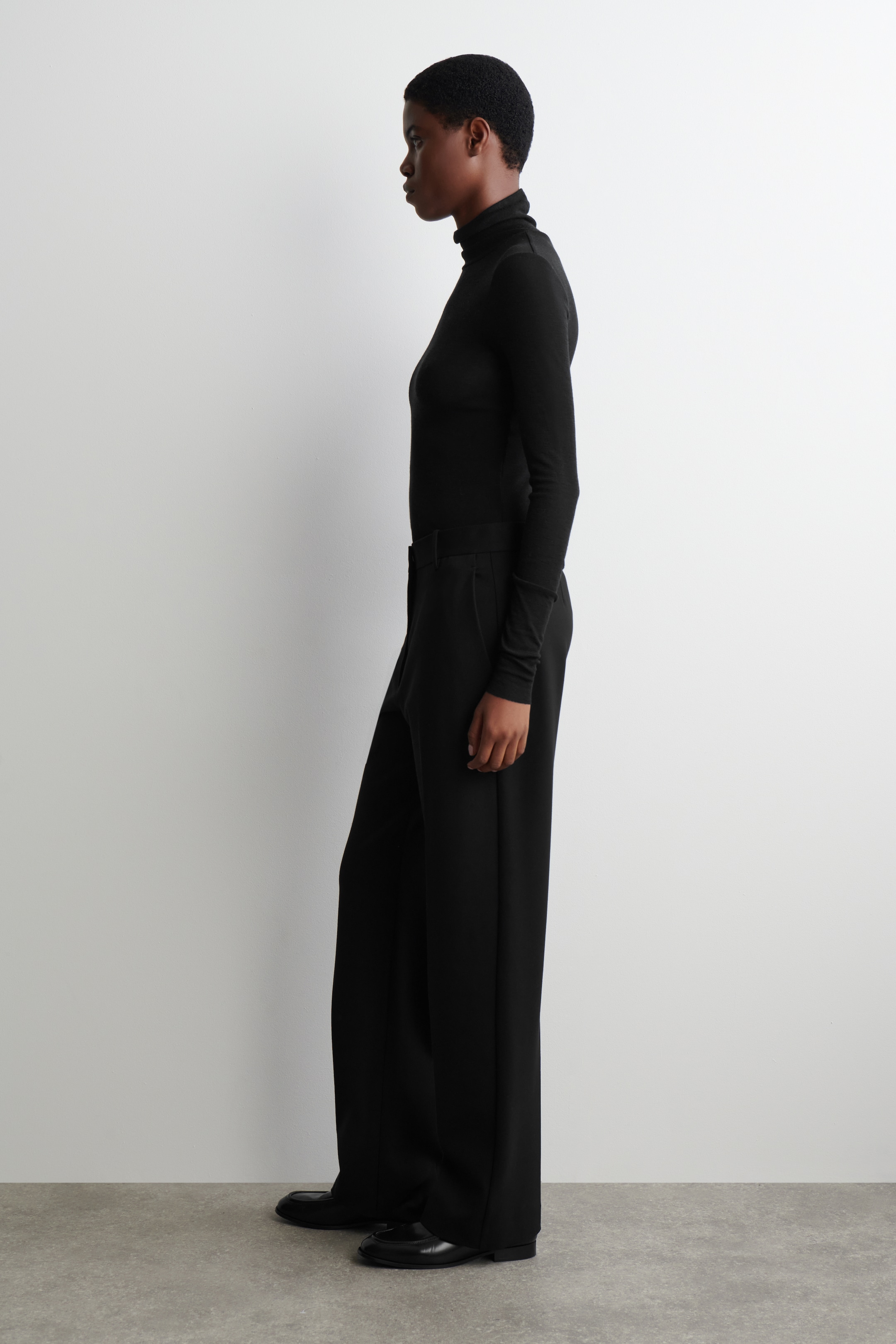 Agrandir l'image: Un haut à col roulé noir, ajusté et à manches longues est porté, rentré dans un pantalon noir à jambes larges. Le pantalon tombe sur des chaussures plates noires, les produits étant montrés de profil.