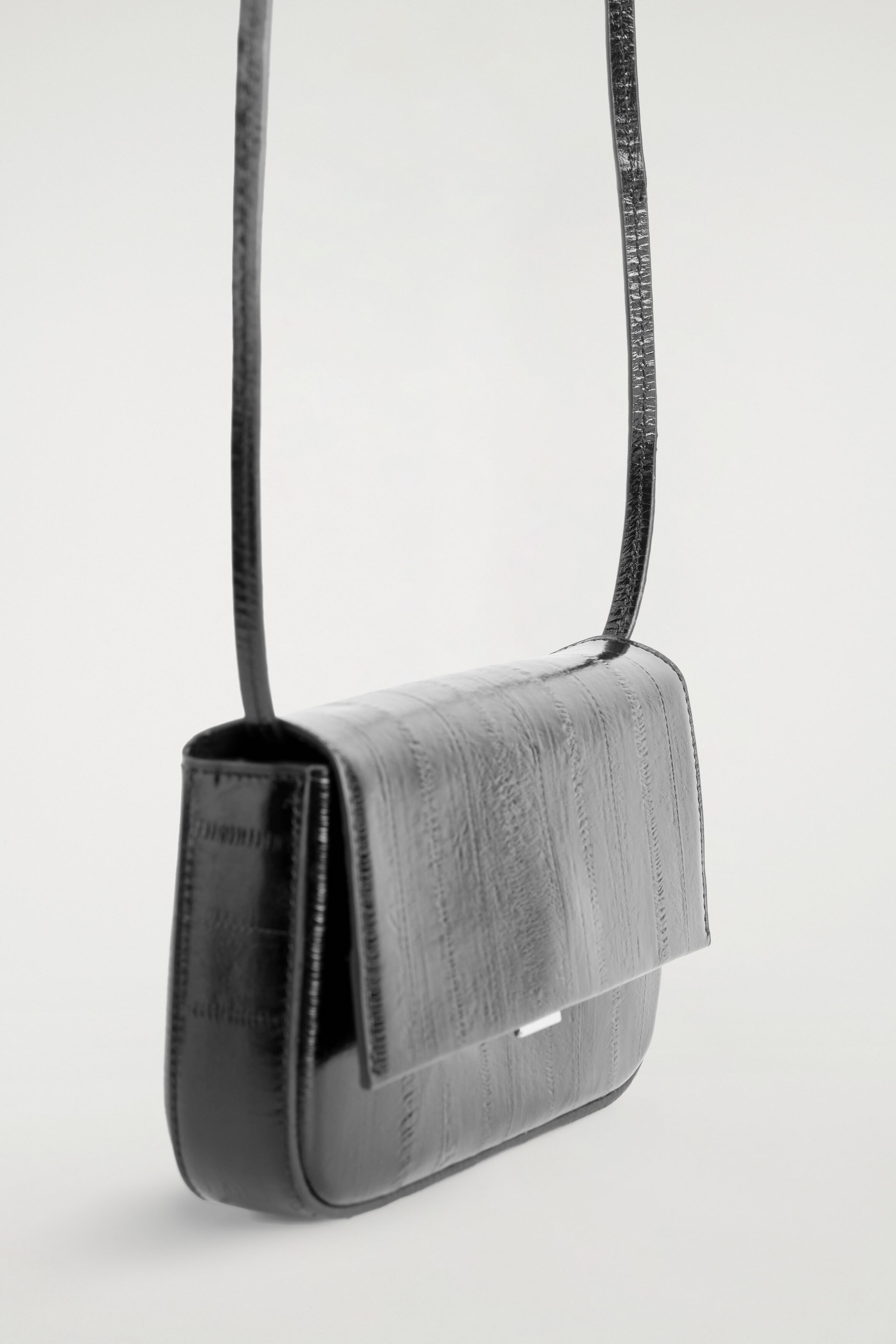 The black eel-effect leather shoulder bag is hanging suspended at a three-quarter front angle, showcasing its structured shape and glossy, vertically panelled texture. A thin leather shoulder strap is extending upwards, while a small silver-tone clasp is peeking out from underneath the front flap. The black eel-effect leather shoulder bag is hanging suspended at a three-quarter front angle, showcasing its structured shape and glossy, vertically panelled texture. A thin leather shoulder strap is extending upwards, while a small silver-tone clasp is peeking out from underneath the front flap.