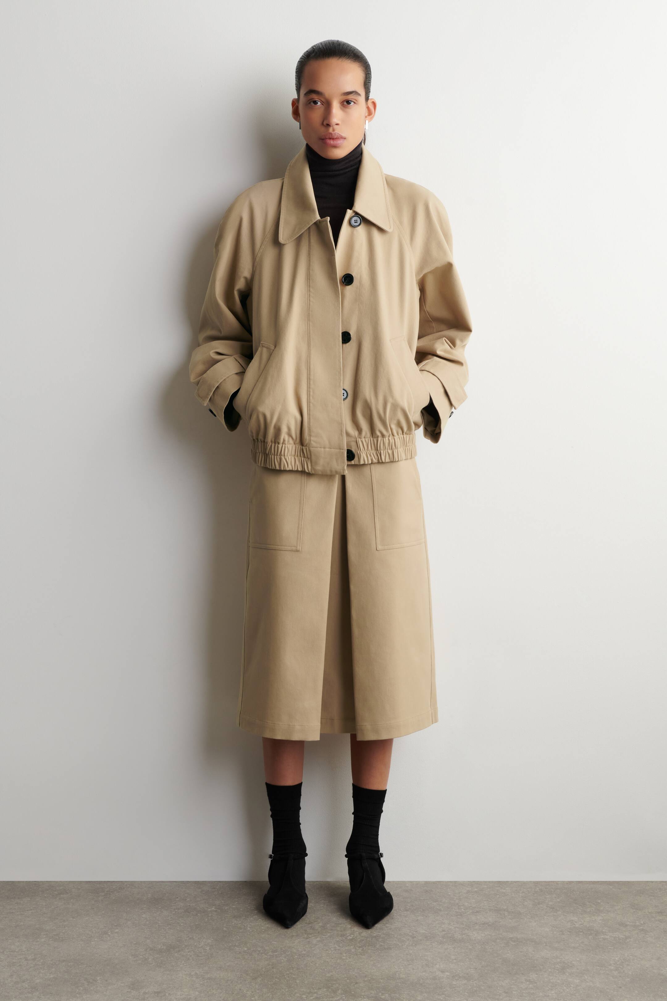 Zobacz większy obraz: Beżowa użytkowa kurtka oraz dopasowana do niej spódnica o długości midi są prezentowane od PRZODU. Kurtka posiada szpiczasty kołnierz typu spread, długie rękawy, marszczony elastyczny dół oraz zapięcie z przodu na czarne guziki, natomiast spódnica została zaprojektowana z zakładkami z przodu i dwiema naszywanymi kieszeniami.