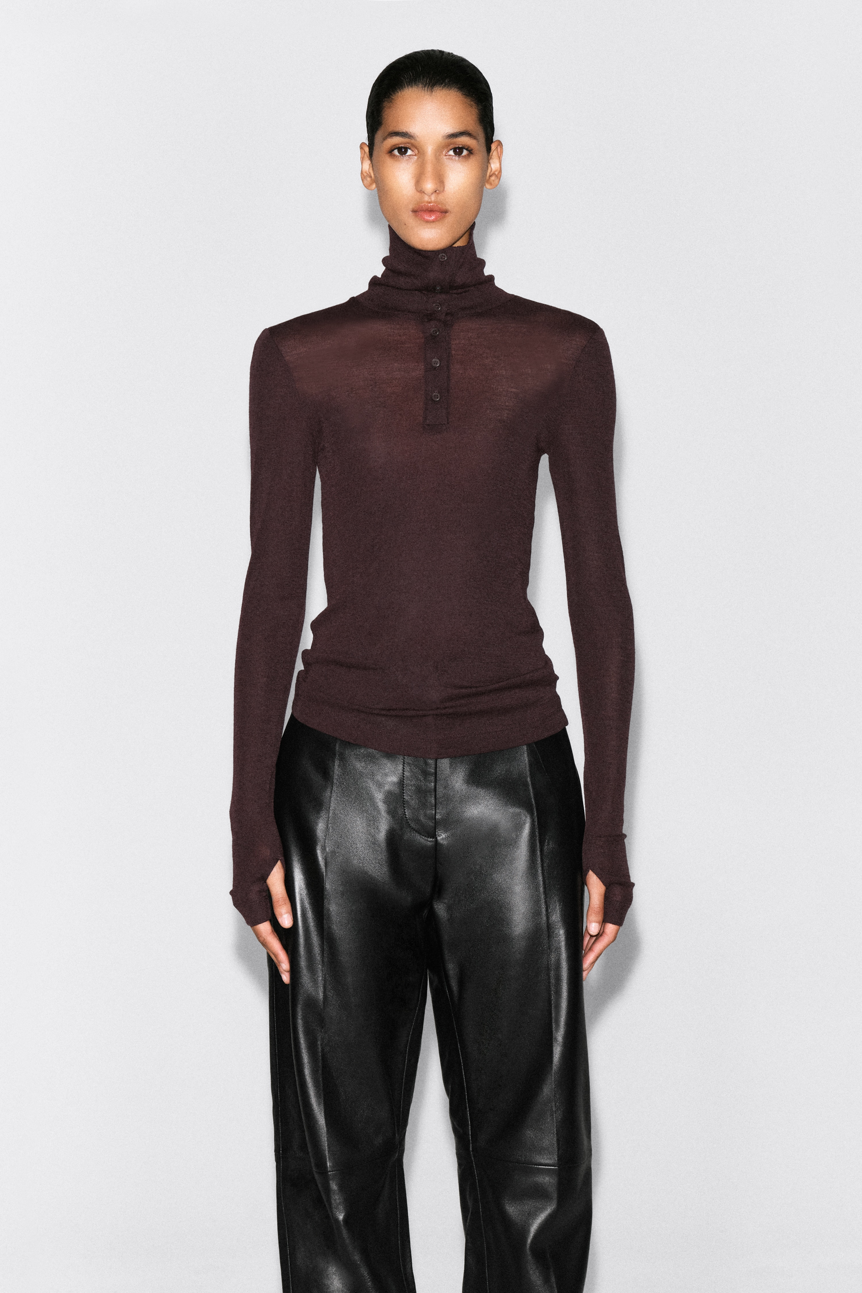 Vista frontale del top con collo a imbuto abbottonato in lana merino, in una tonalitÃ  marrone prugna scuro, caratterizzato da una silhouette slim fit e maniche lunghe con sottili spacchi ai polsini. Il top mostra unâabbottonatura parziale sul davanti e un colletto convertibile a collo a imbuto con asole orizzontali.