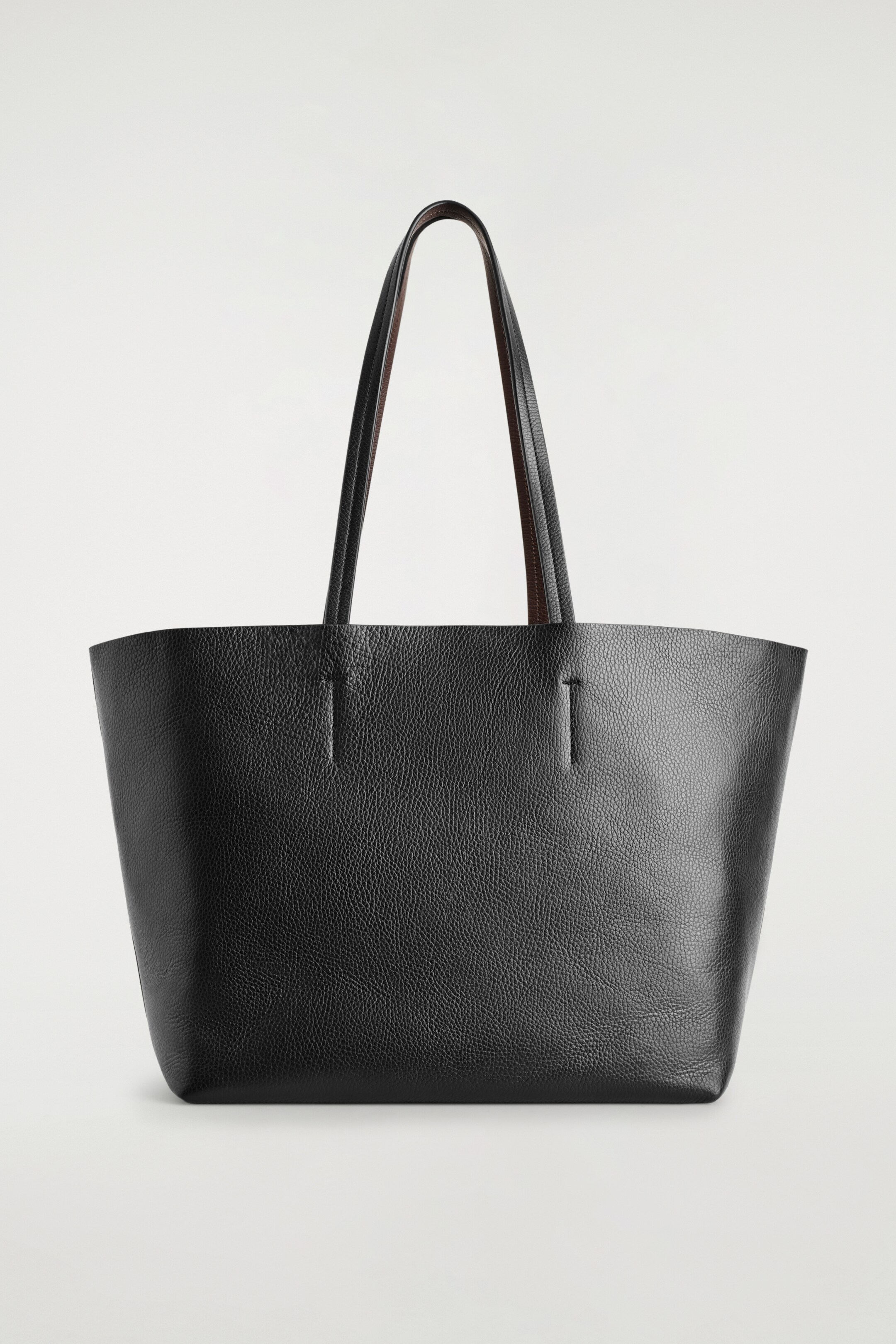 Agrandir l'image: Un sac fourre-tout en cuir noir grainé est orienté FACE avant, avec deux poignées fines arquées vers le haut à partir de fentes verticales sur son corps. Les dessous des poignées apparaissent dans une couleur marron contrastante.