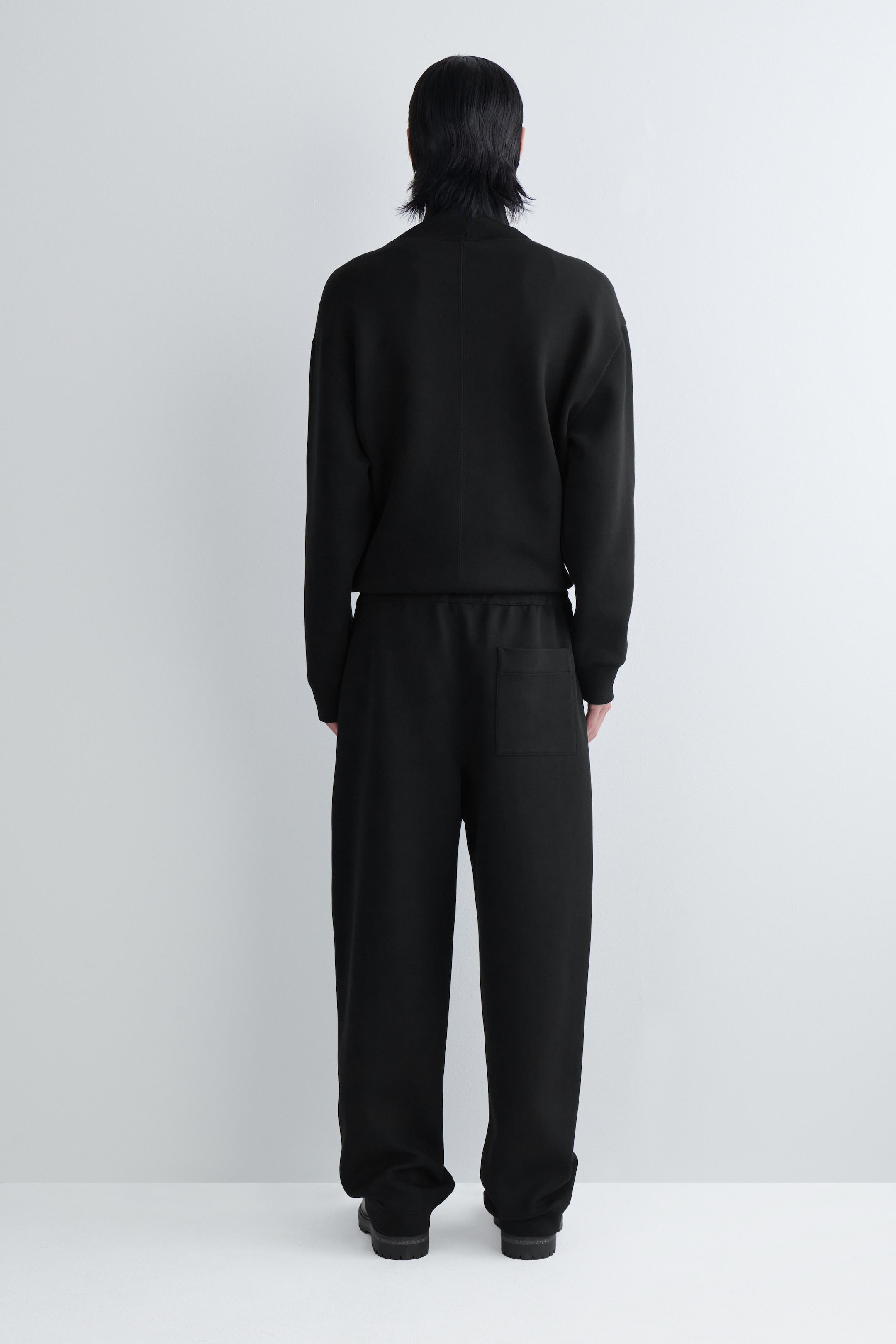 Agrandir l'image: Un haut noir à manches longues et col montant ainsi qu'un pantalon assorti à coupe ample sont présentés de dos, montrant une coupe décontractée. Le haut présente une couture centrale au dos, et le pantalon affiche une taille élastique, une seule poche arrière plaquée et une coupe large, avec des chaussures noires complétant le look.