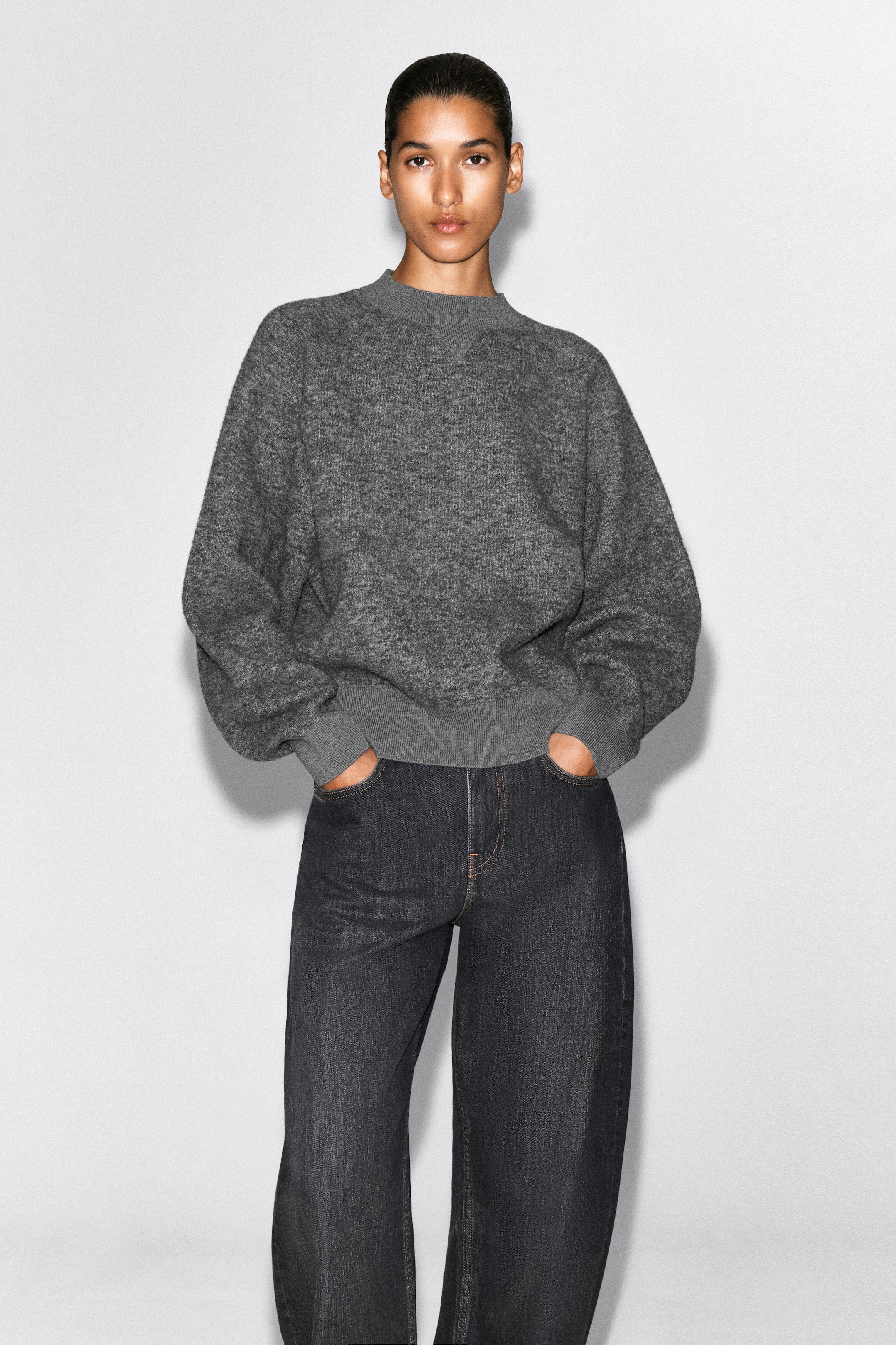 This heather gray knit sweater showcases a relaxed, slightly oversized fit with a ribbed mock neck featuring a subtle triangular stitch detail at the collar. The sweater has voluminous long sleeves with wide ribbed cuffs and a matching ribbed hem resting at the waist of dark wash jeans. This heather gray knit sweater showcases a relaxed, slightly oversized fit with a ribbed mock neck featuring a subtle triangular stitch detail at the collar. The sweater has voluminous long sleeves with wide ribbed cuffs and a matching ribbed hem resting at the waist of dark wash jeans.