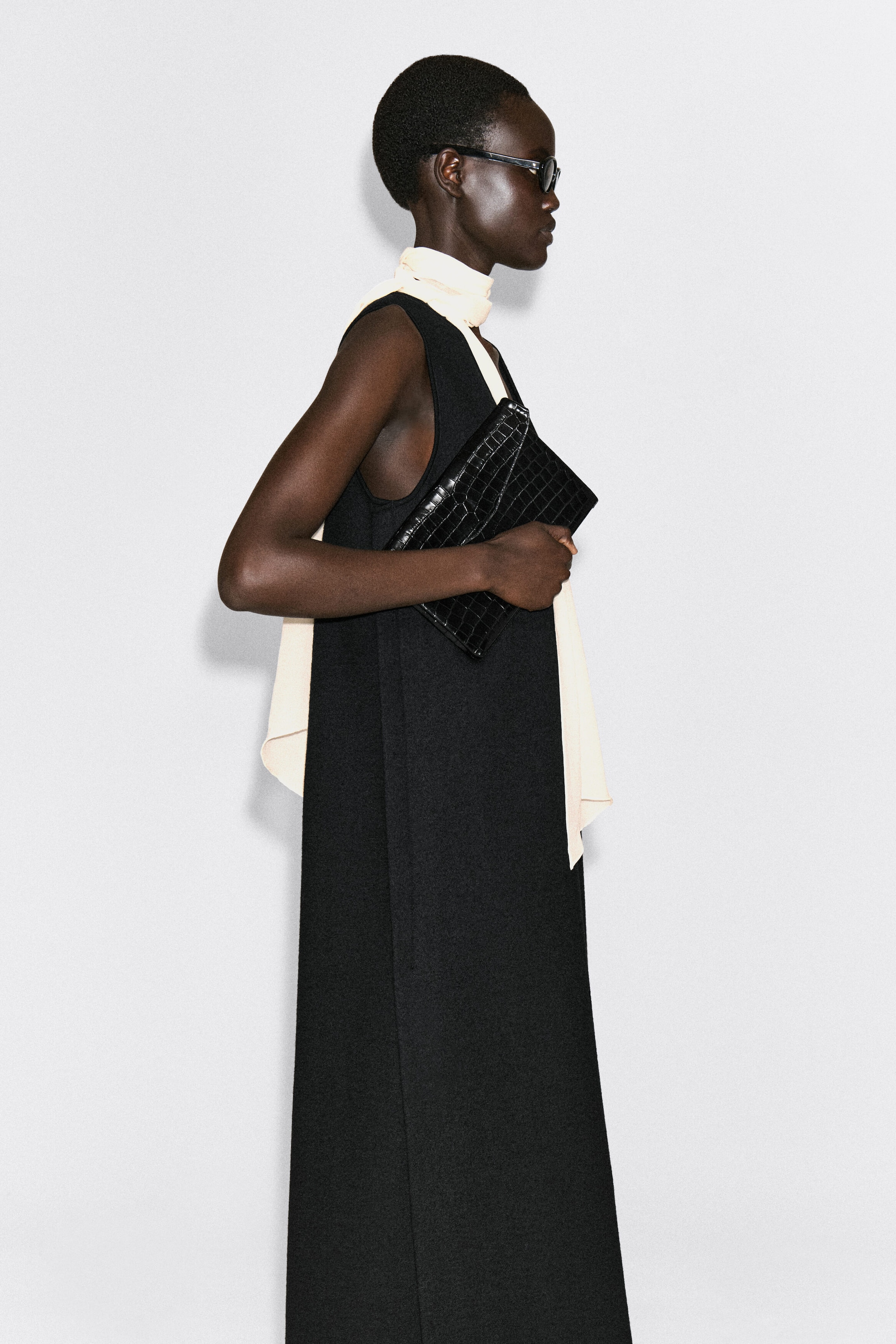 The black croc-embossed leather Envelope Clasp Pouch is being held by a model facing right, showcasing its glossy, textured finish and structured rectangular shape. The pouch features a triangular envelope-style flap and is styled tucked under the arm to demonstrate its scale and design. The black croc-embossed leather Envelope Clasp Pouch is being held by a model facing right, showcasing its glossy, textured finish and structured rectangular shape. The pouch features a triangular envelope-style flap and is styled tucked under the arm to demonstrate its scale and design.