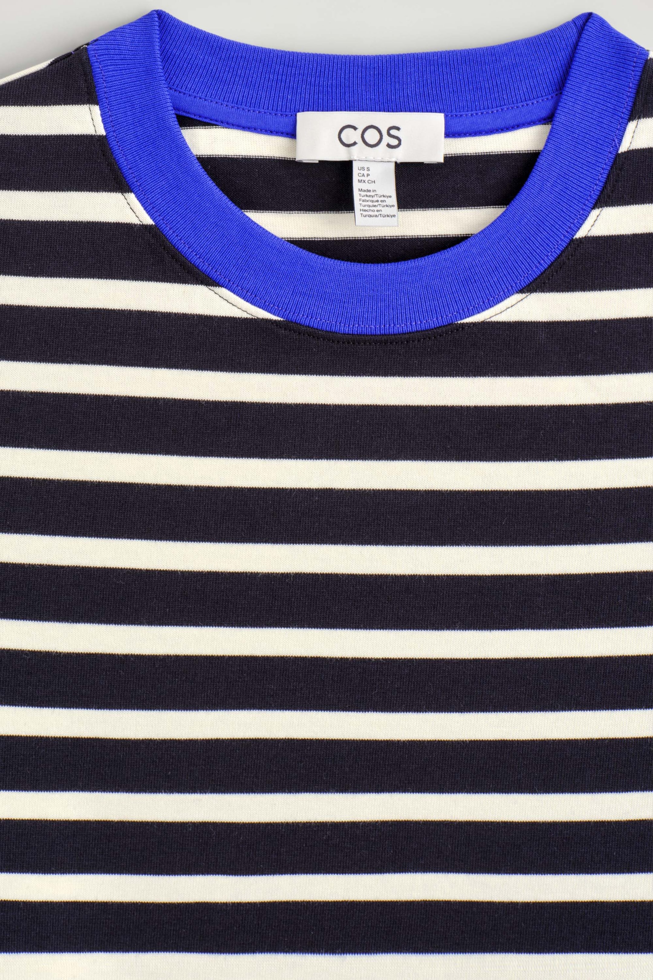 The Clean Cut Long-Sleeved T-Shirt showcases a black and off-white horizontal stripe pattern in a close-up front view. The garment features a contrasting bright blue ribbed crew neck, with a white brand label visible on the inside back collar. The Clean Cut Long-Sleeved T-Shirt showcases a black and off-white horizontal stripe pattern in a close-up front view. The garment features a contrasting bright blue ribbed crew neck, with a white brand label visible on the inside back collar.