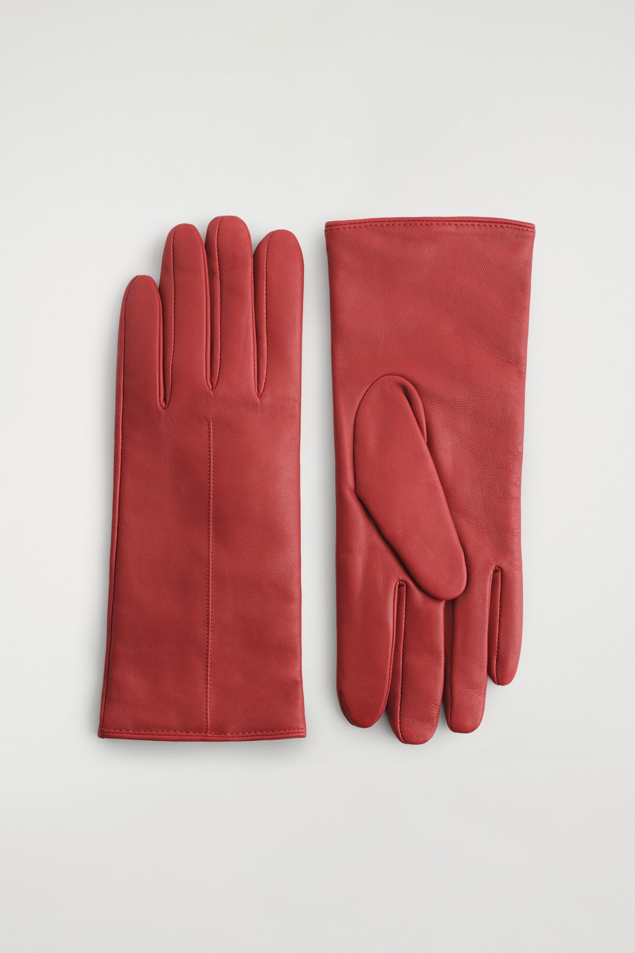 Une paire de gants en cuir rouge est disposée à plat, l’un montrant le dos de la main avec une couture verticale apparente, et l’autre montrant la construction du côté paume. Les deux gants présentent une texture de cuir lisse avec de fines surpiqûres et des ourlets cousus nets aux poignets.