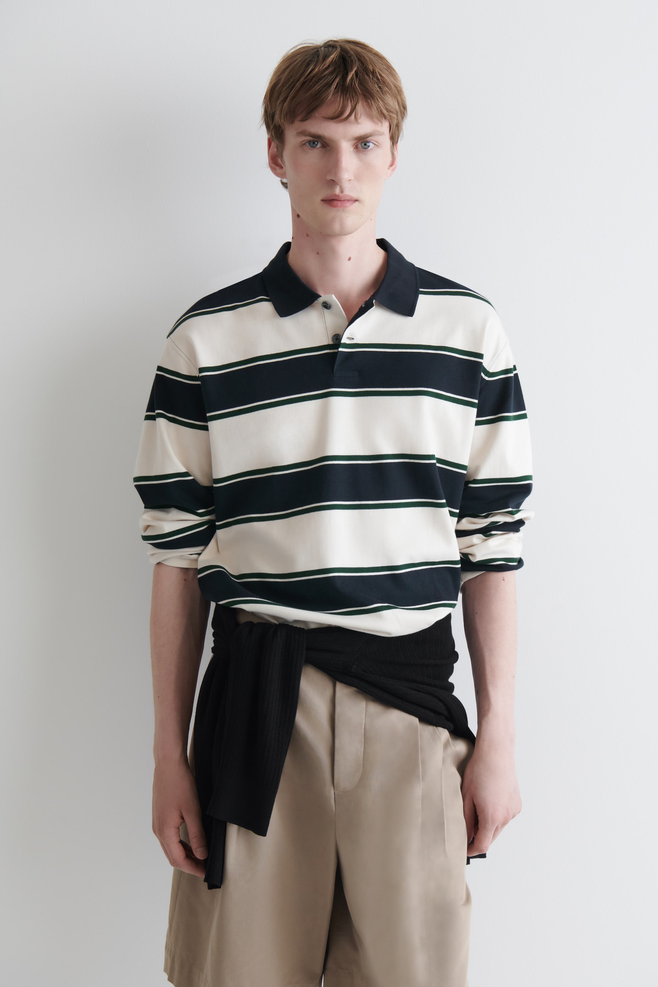 Agrandir l'image: Un polo à manches longues à coupe décontractée, de couleur crème avec des rayures horizontales bleu marine foncé, chacune rehaussée d’une fine ligne verte, est porté par le mannequin. Un pull noir est noué autour de la taille du mannequin, par-dessus le polo.