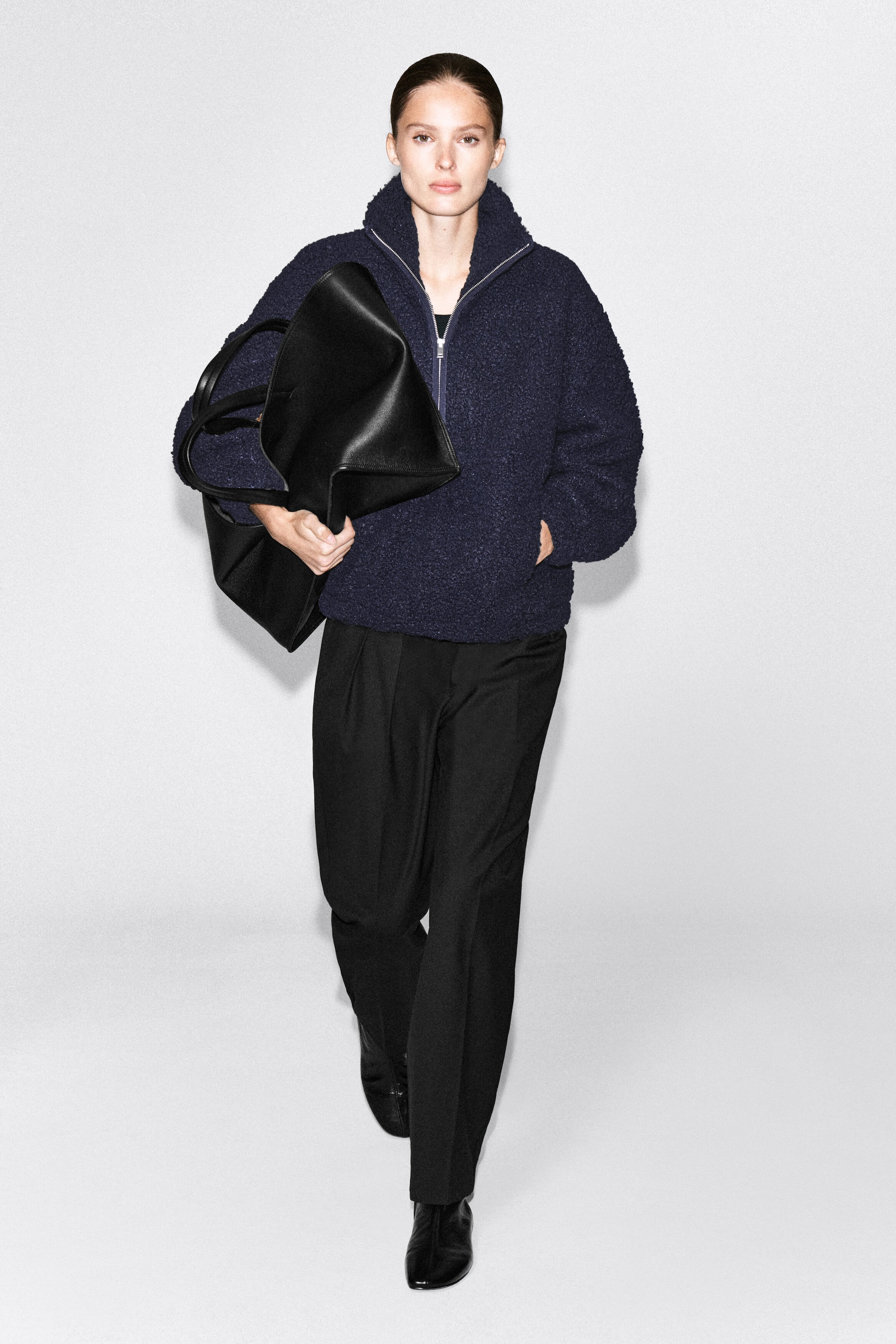 A dark blue, textured teddy half-zip jumper is being worn by the model, who is walking with one hand in its side pocket. The jumper features a funnel neck with a partially open zipper and is styled with black trousers and a large black leather tote bag. A dark blue, textured teddy half-zip jumper is being worn by the model, who is walking with one hand in its side pocket. The jumper features a funnel neck with a partially open zipper and is styled with black trousers and a large black leather tote bag.