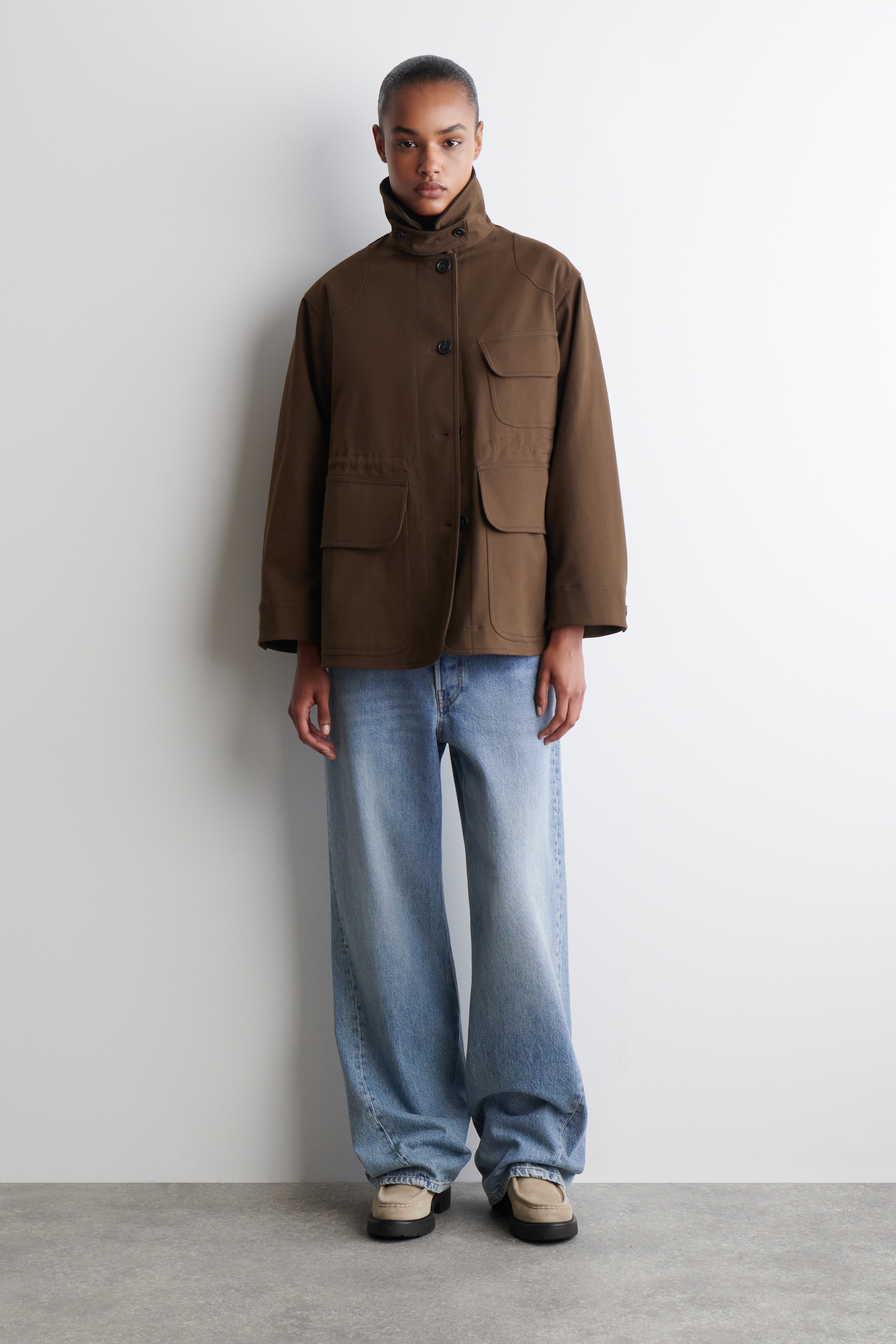 Agrandir l'image: Une veste utilitaire brun foncé est présentée de face, mettant en valeur son design structuré. Elle est dotée d'un col montant, d'une patte de boutonnage visible à l'avant et de quatre grandes poches à rabat, deux sur la poitrine et deux sur le bas avant. Un cordon de serrage subtil resserre la taille.