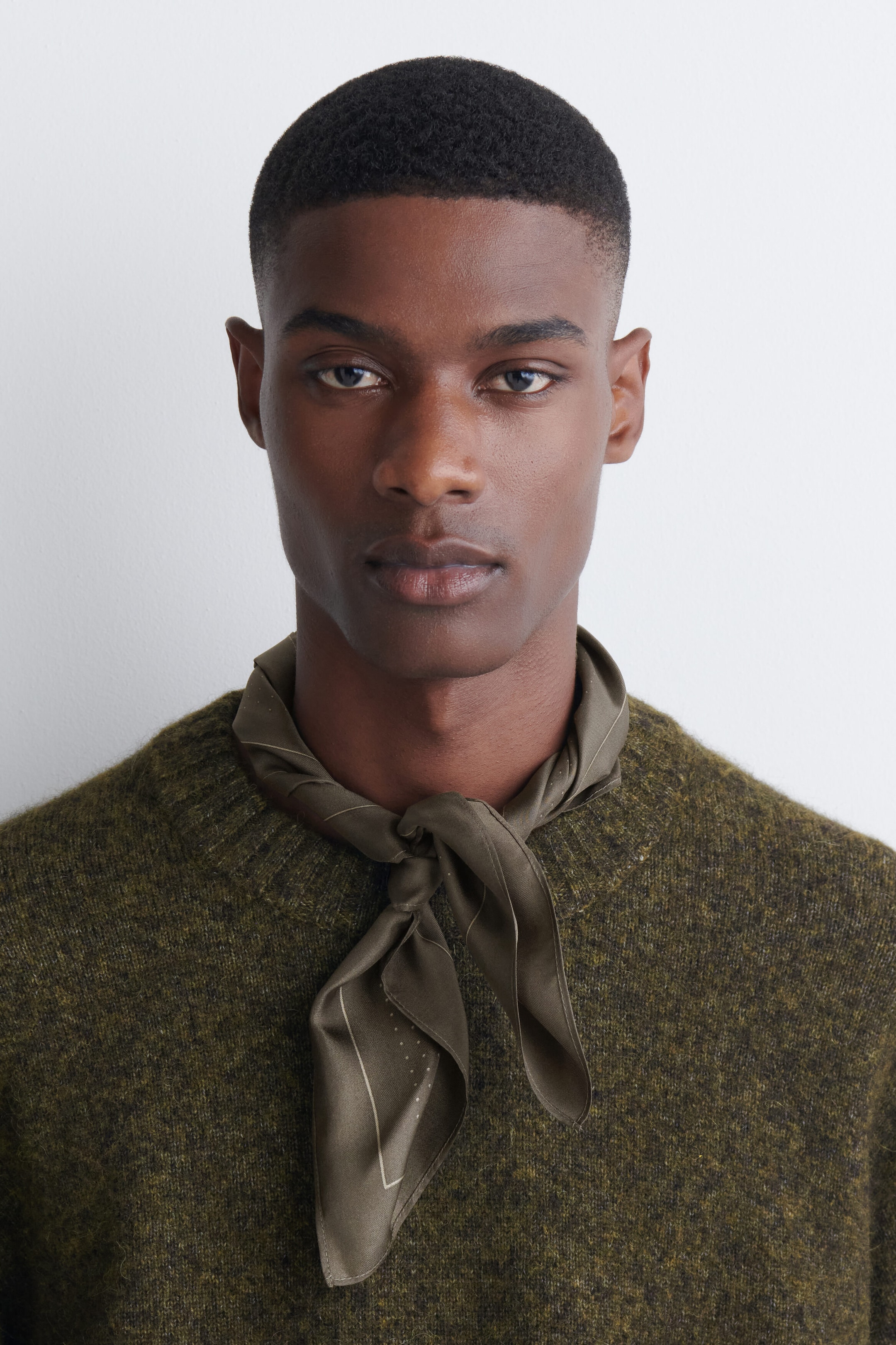 A polka-dot silk pocket square in olive green is being worn as a neckerchief, tied in a knot around the model’s neck. The accessory features a delicate dot pattern and border, draping over a textured olive-green knit sweater. A polka-dot silk pocket square in olive green is being worn as a neckerchief, tied in a knot around the model’s neck. The accessory features a delicate dot pattern and border, draping over a textured olive-green knit sweater.
