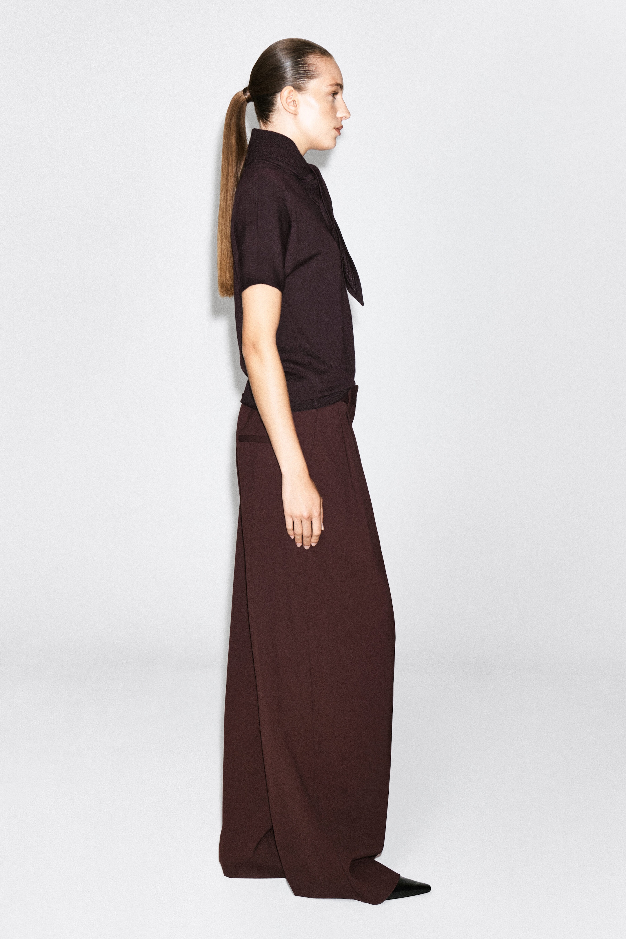 The model is wearing a dark brown merino wool-blend T-shirt with scarf detailing, featuring short sleeves and a high collar with an integrated draped scarf. The T-shirt is styled tucked into coordinating wide-leg trousers.