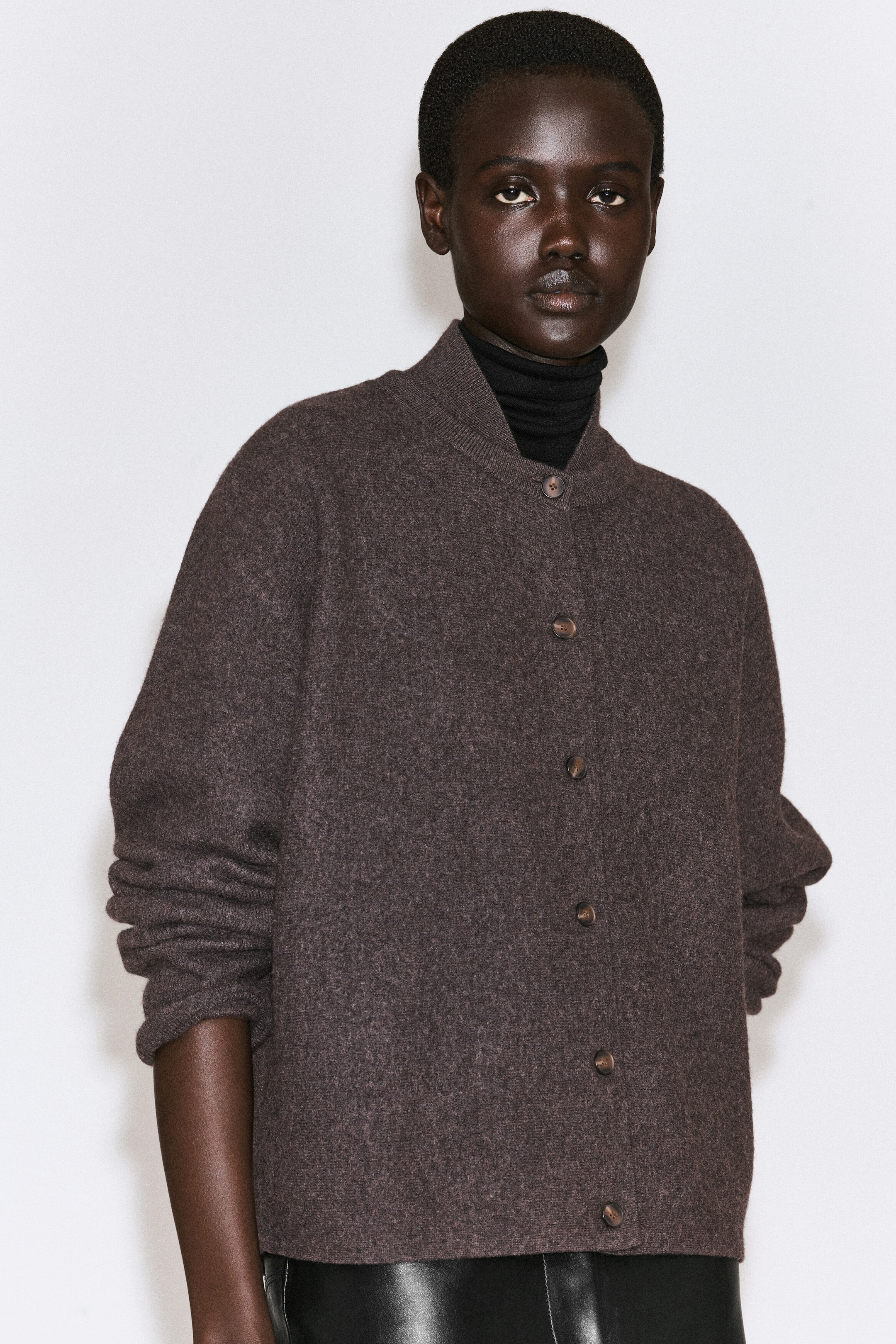 Un cardigan en laine bouillie brun foncé présente une silhouette doucement arrondie, un col côtelé de style bomber et une fermeture boutonnée sur le devant. Le cardigan est porté en superposition sur un col roulé noir, mettant en valeur sa coupe décontractée et son tissu texturé. Un cardigan en laine bouillie brun foncé présente une silhouette doucement arrondie, un col côtelé de style bomber et une fermeture boutonnée sur le devant. Le cardigan est porté en superposition sur un col roulé noir, mettant en valeur sa coupe décontractée et son tissu texturé.