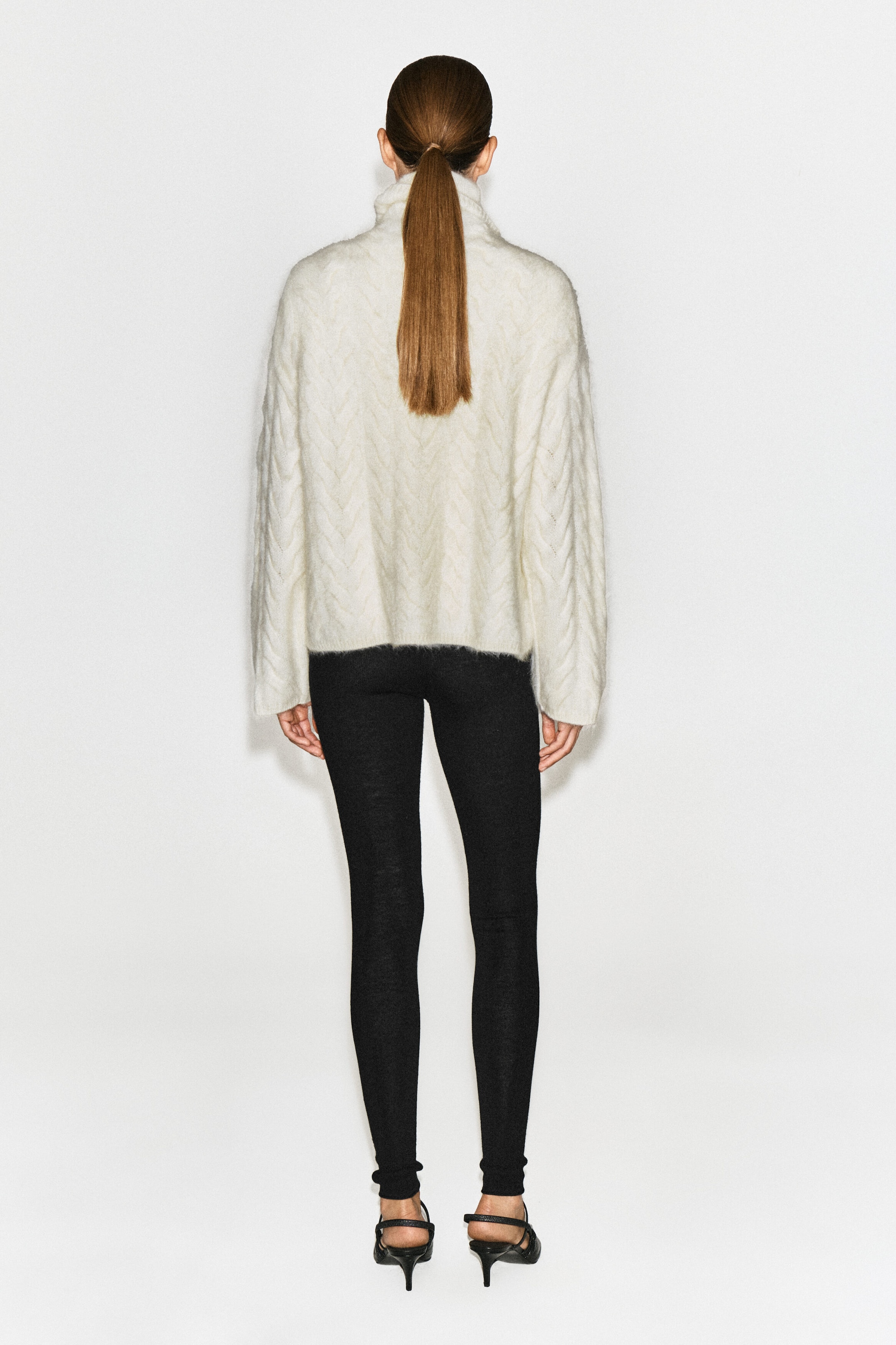 A cable-knit brushed cashmere funnel-neck sweater in an off-white shade is worn by the model, shown from the back. The sweater features a fuzzy, textured knit, long sleeves, and a relaxed fit, and is styled with black leggings and black heels.