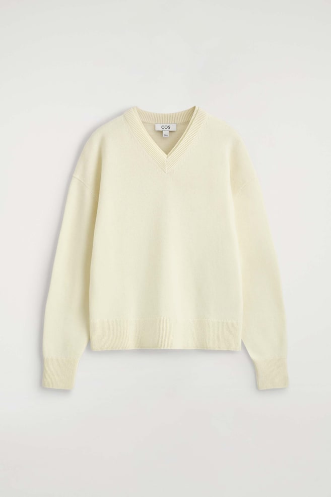 Merino Wool Layered V-Neck Jumper vises forfra i en kremfarget strikket design med en avslappet passform. Den har en dobbel lagdelt ribbestrikket V-hals, senkede skuldre, lange ermer med ribbestrikkede mansjetter og en matchende ribbestrikket kant. - 1