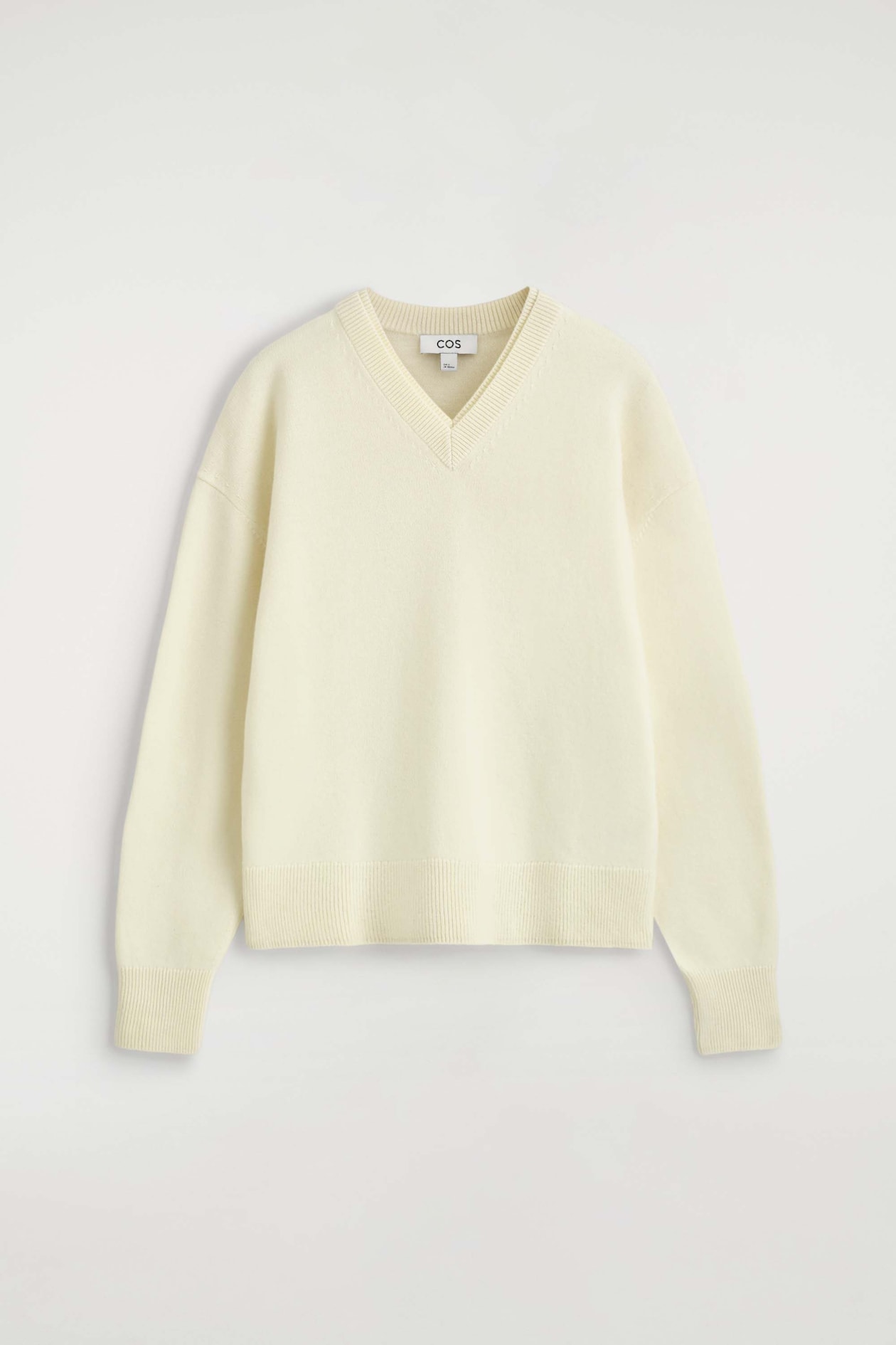 The Merino Wool Layered V-Neck Jumper is shown from the front in a cream-coloured knit design with a relaxed fit. It features a double-layered ribbed V-neckline, dropped shoulders, long sleeves with ribbed cuffs, and a matching ribbed hem.