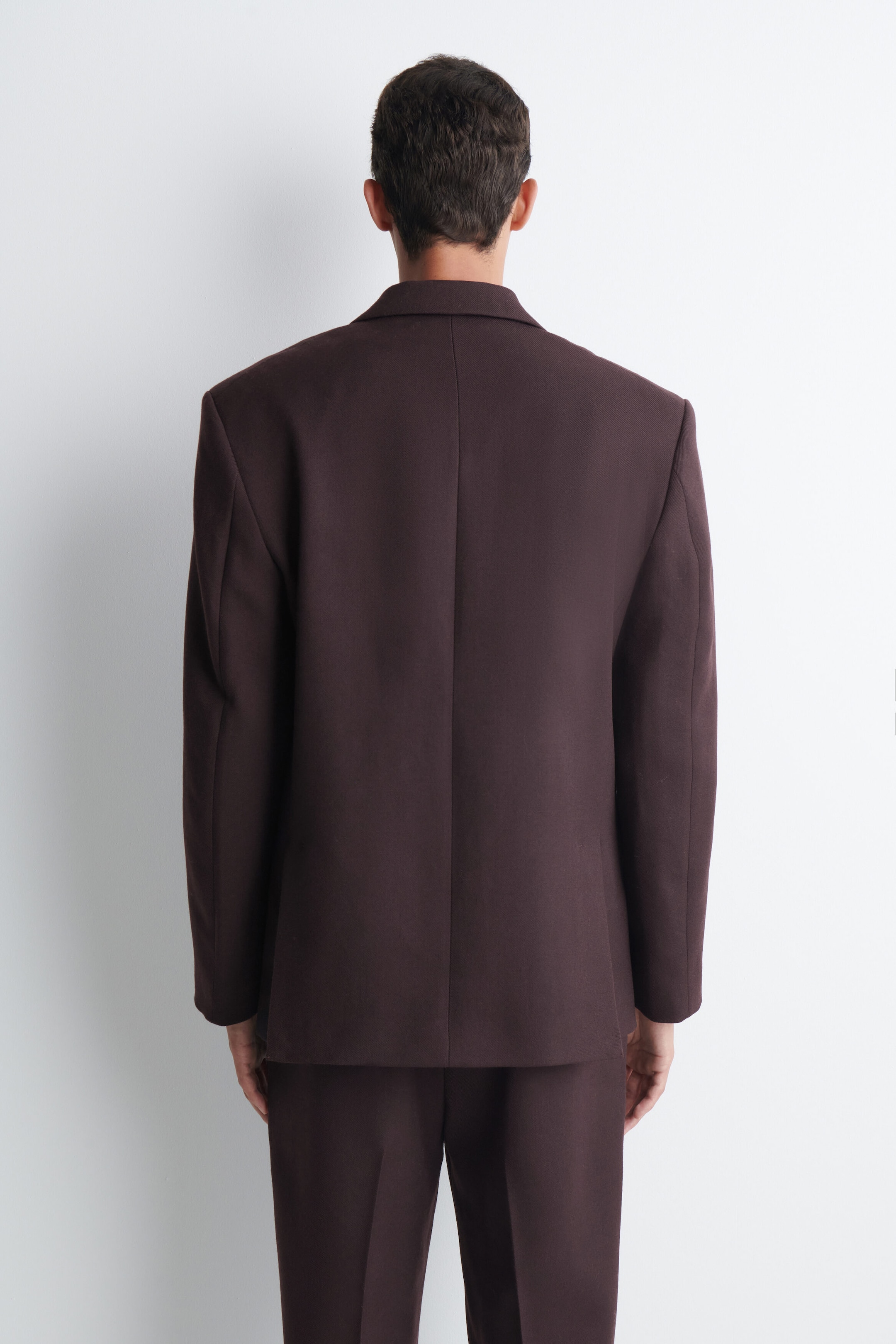 The model is wearing a dark brown suit comprising a blazer and trousers. The blazer features a structured back and long sleeves. The model is wearing a dark brown suit comprising a blazer and trousers. The blazer features a structured back and long sleeves.