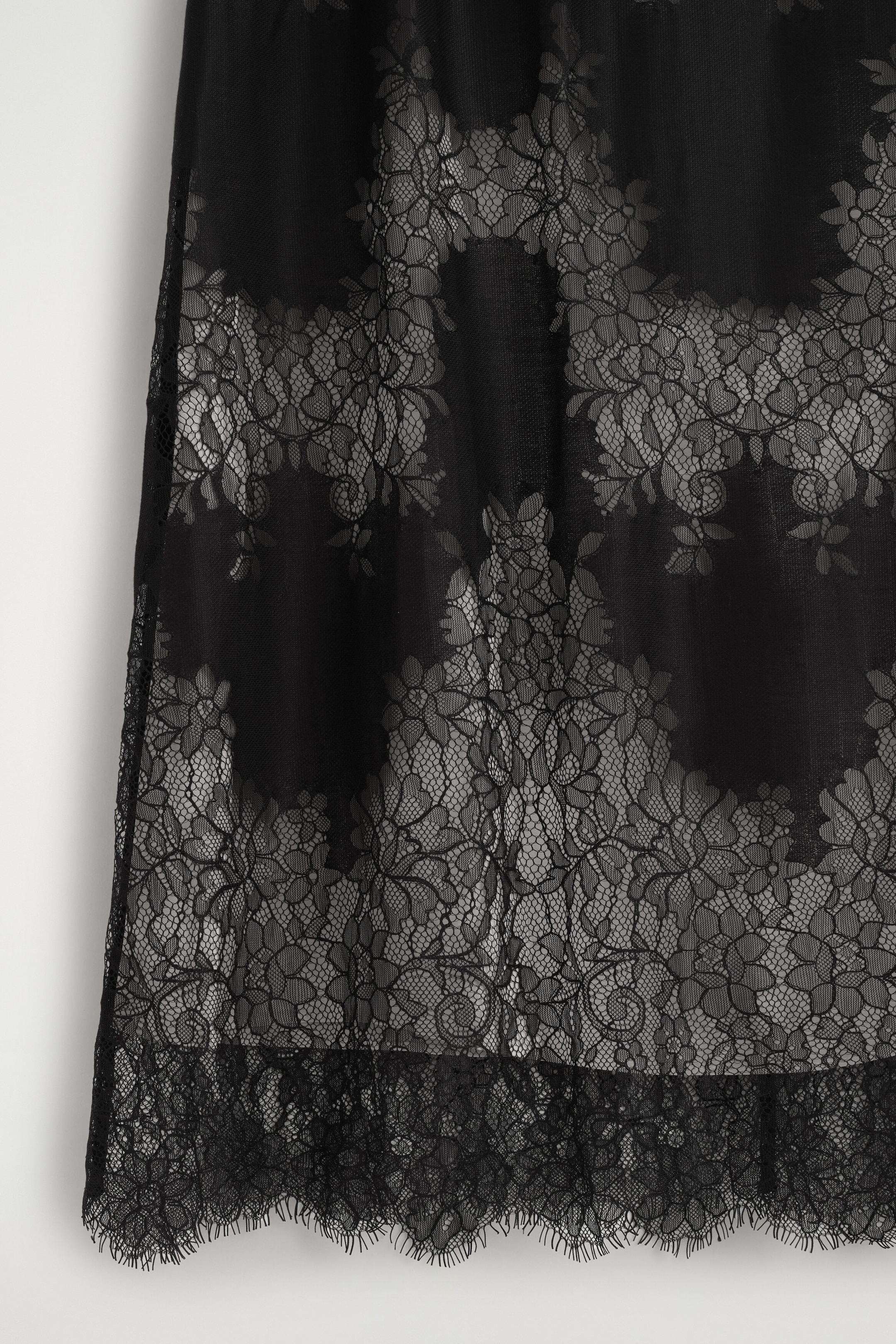 A sleeveless lace midi dress is shown from the front, highlighting the lower skirt, with a sheer black floral lace overlay. The lace overlay extends beyond a shorter, solid black inner lining and finishes with a scalloped eyelash lace hem. A sleeveless lace midi dress is shown from the front, highlighting the lower skirt, with a sheer black floral lace overlay. The lace overlay extends beyond a shorter, solid black inner lining and finishes with a scalloped eyelash lace hem.
