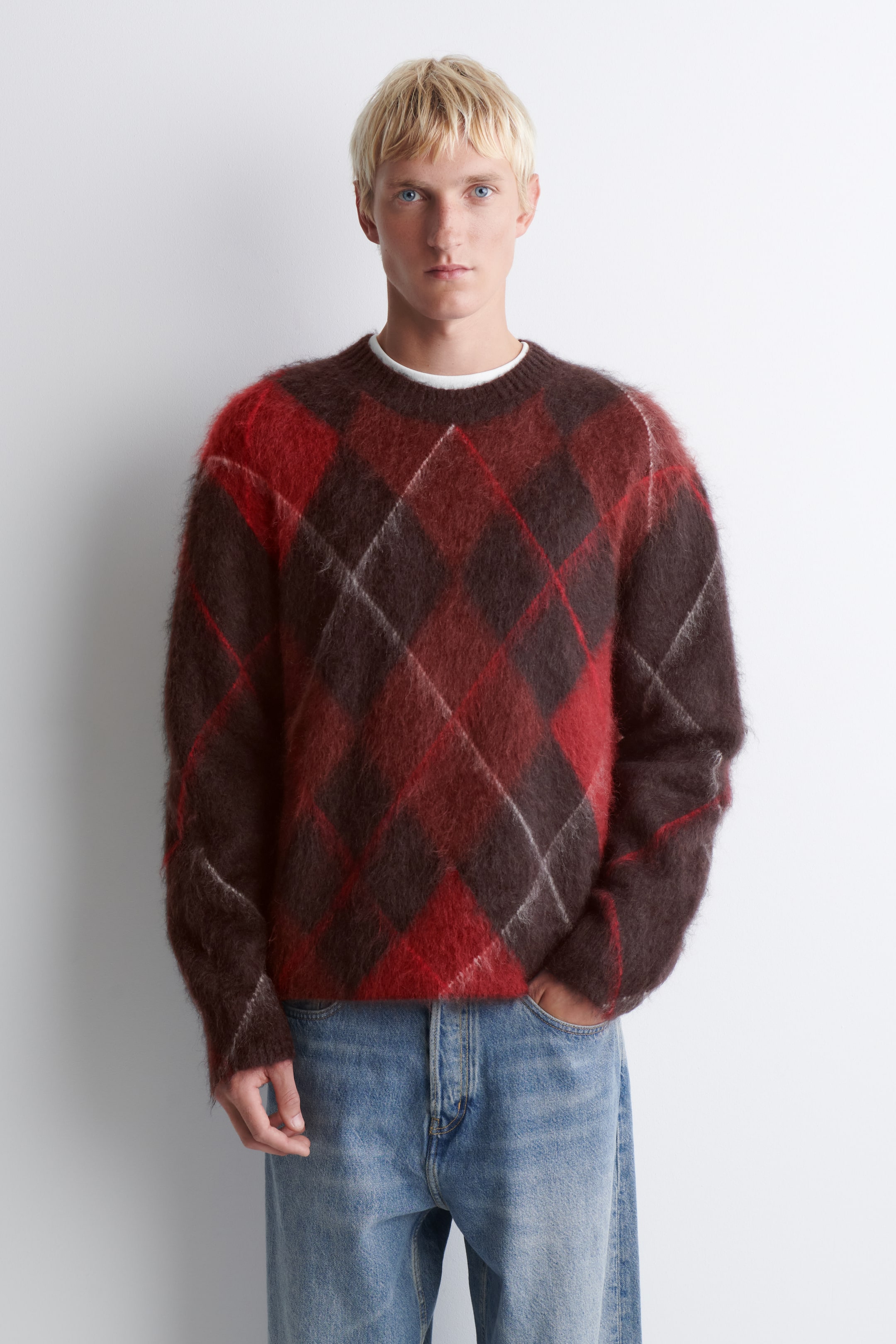 View larger image: The crew neck sweater is shown from the front, featuring a fuzzy, brushed-knit texture with a large red and dark brown argyle pattern accented by thin white lines. It has a relaxed fit with long sleeves and is styled over a visible white T-shirt collar.