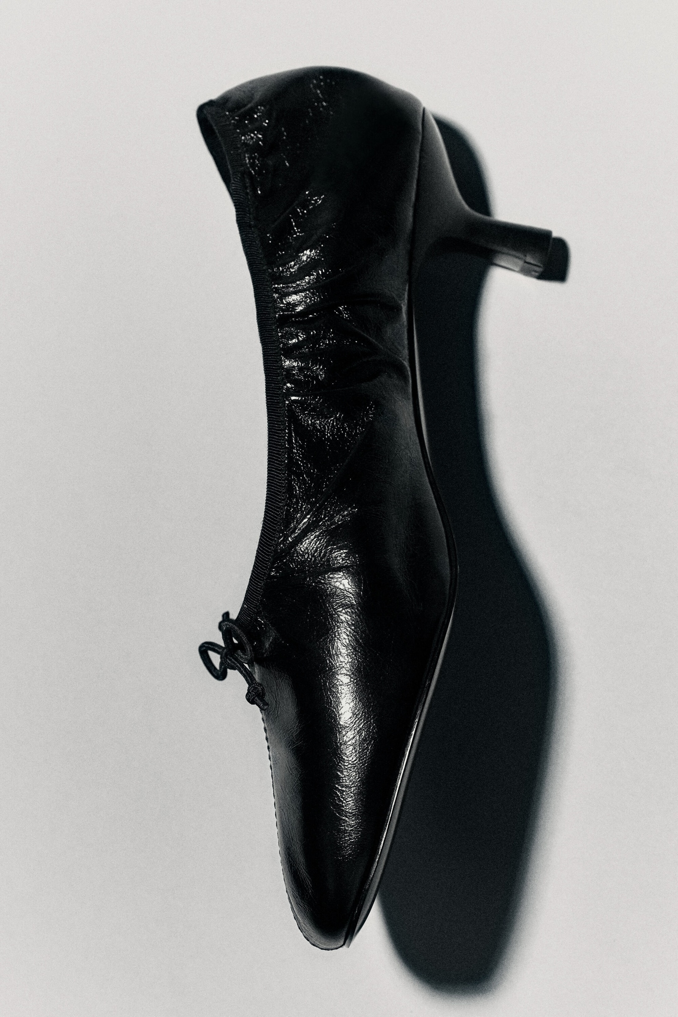 A black leather heeled ballet pump is displaying a glossy, crinkled texture and a low kitten heel, presented from a side profile. The shoe features a tapered toe, a small bow detail at the front, and a gathered, elasticated trim along the opening. A black leather heeled ballet pump is displaying a glossy, crinkled texture and a low kitten heel, presented from a side profile. The shoe features a tapered toe, a small bow detail at the front, and a gathered, elasticated trim along the opening.