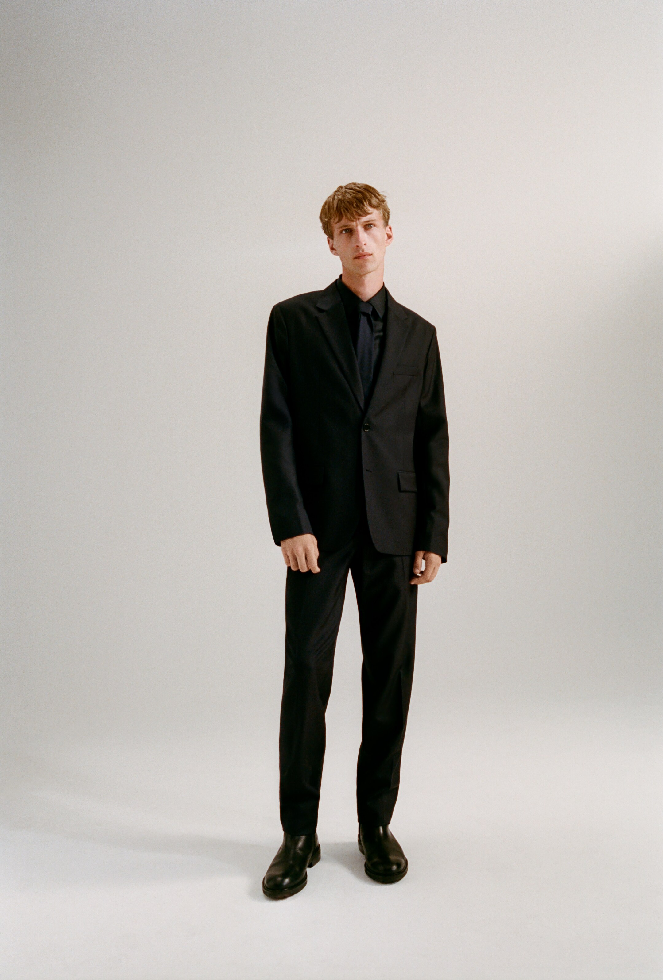 Größeres Bild anzeigen: Ein schwarzer einreihiger Anzug wird von vorne präsentiert, mit einem Blazer mit Kerbrevers, geknöpften Manschetten und vorderen Pattentaschen, kombiniert mit einem dunklen Hemd und einer Krawatte. Die dazu passenden gerade geschnittenen Hosen zeigen einen sauberen Bund und gebügelte Falten an der Vorderseite.