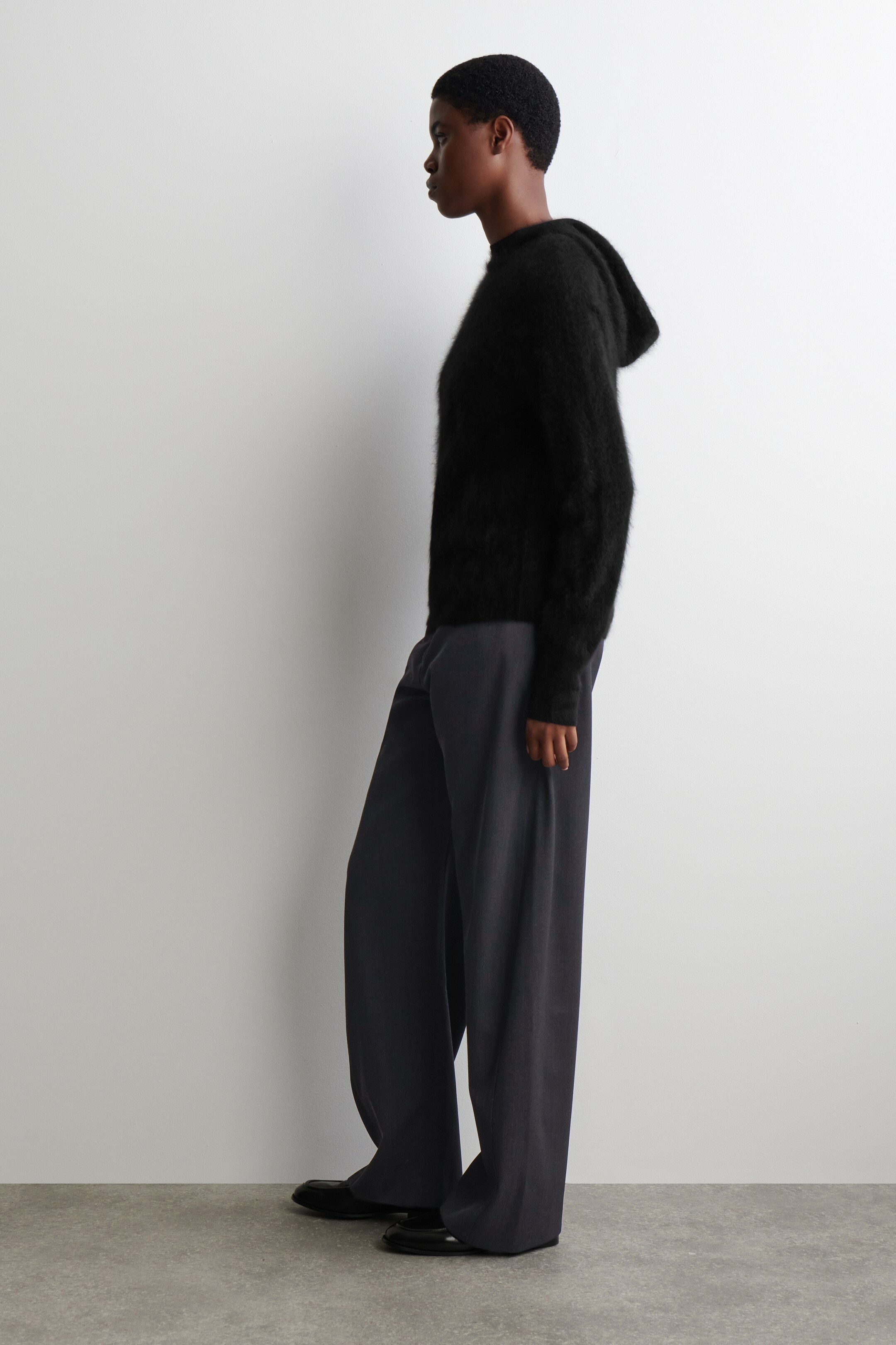 View larger image: A black fuzzy hooded sweater is being worn with dark grey wide-leg trousers and black flat shoes, all seen from a side profile.