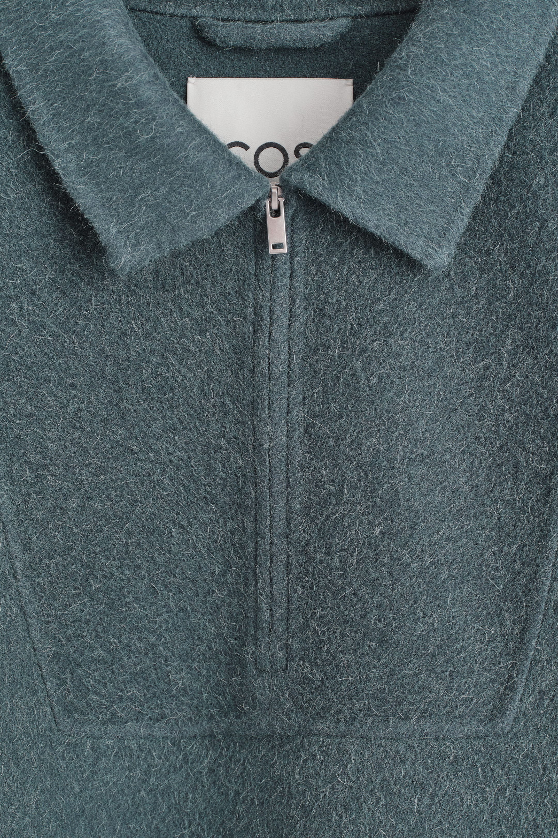 A teal-blue, fuzzy textured knit sweater is shown in a close-up front view, featuring a pointed polo collar and a silver-tone half-zip closure. The design includes a rectangular stitched panel framing the zipper and a partially visible white brand label inside the collar. A teal-blue, fuzzy textured knit sweater is shown in a close-up front view, featuring a pointed polo collar and a silver-tone half-zip closure. The design includes a rectangular stitched panel framing the zipper and a partially visible white brand label inside the collar.