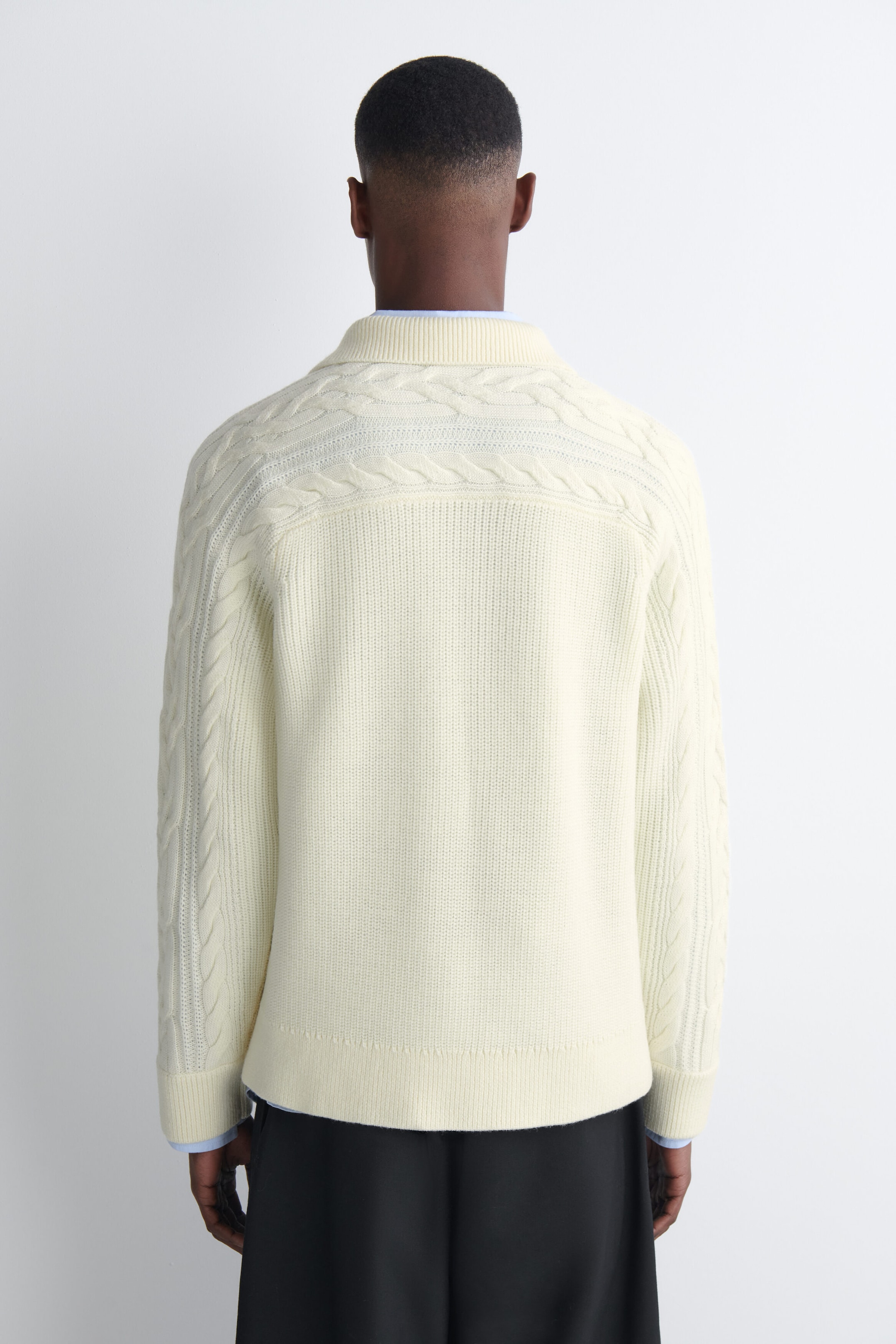 A cream-colored chunky knit polo sweater featuring cable-knit detailing across the shoulders and sleeves is worn by the model. The sweater is layered over a light blue collared shirt and paired with black trousers. A cream-colored chunky knit polo sweater featuring cable-knit detailing across the shoulders and sleeves is worn by the model. The sweater is layered over a light blue collared shirt and paired with black trousers.