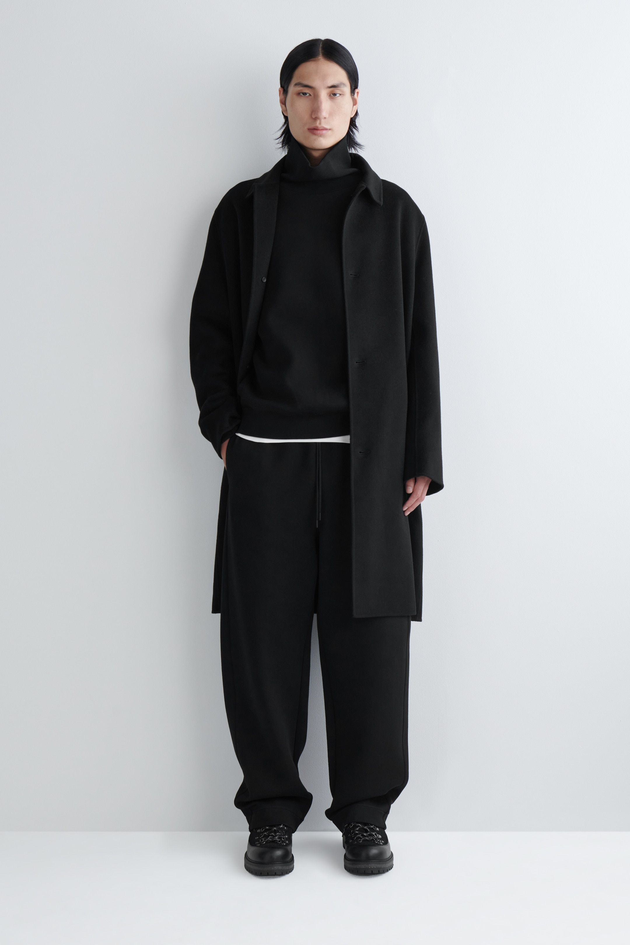 Agrandir l'image: Un manteau noir à coupe décontractée avec un col est porté déboutonné sur un col roulé noir et un pantalon noir à jambes larges avec une taille à cordon visible, sur une personne faisant face vers l'avant. Une couche blanche est visible entre le haut et le pantalon, et des bottes noires à semelles épaisses avec des lacets complètent l'ensemble.