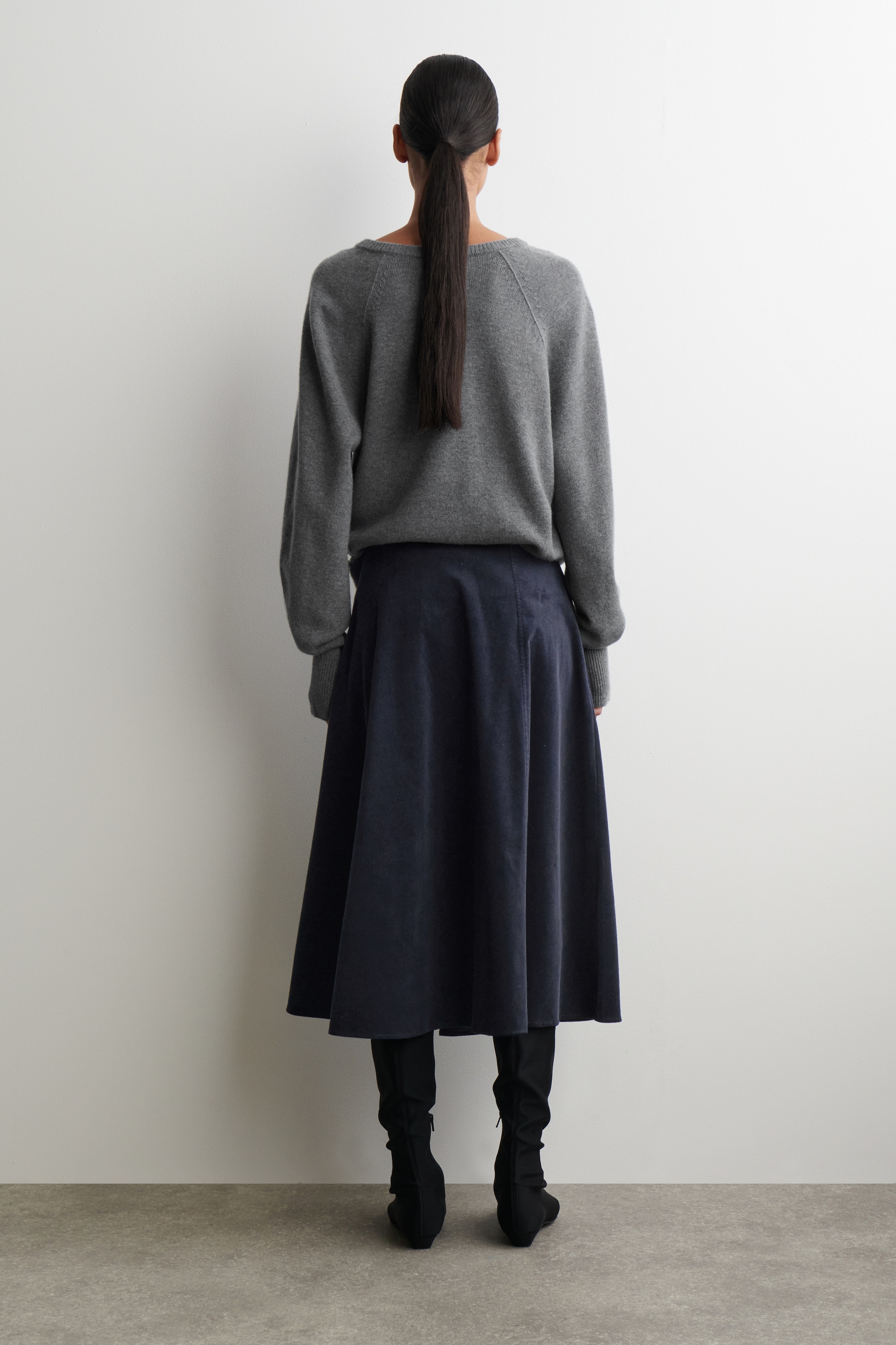Agrandir l'image: Un pull gris à manches raglan, avec des poignets et un ourlet côtelés, est porté, vu de dos. Une jupe midi bleu marine en ligne A est stylée en dessous, montrant une couture arrière visible, tandis que des bottes noires à hauteur de genou avec des talons blocs complètent l'ensemble.