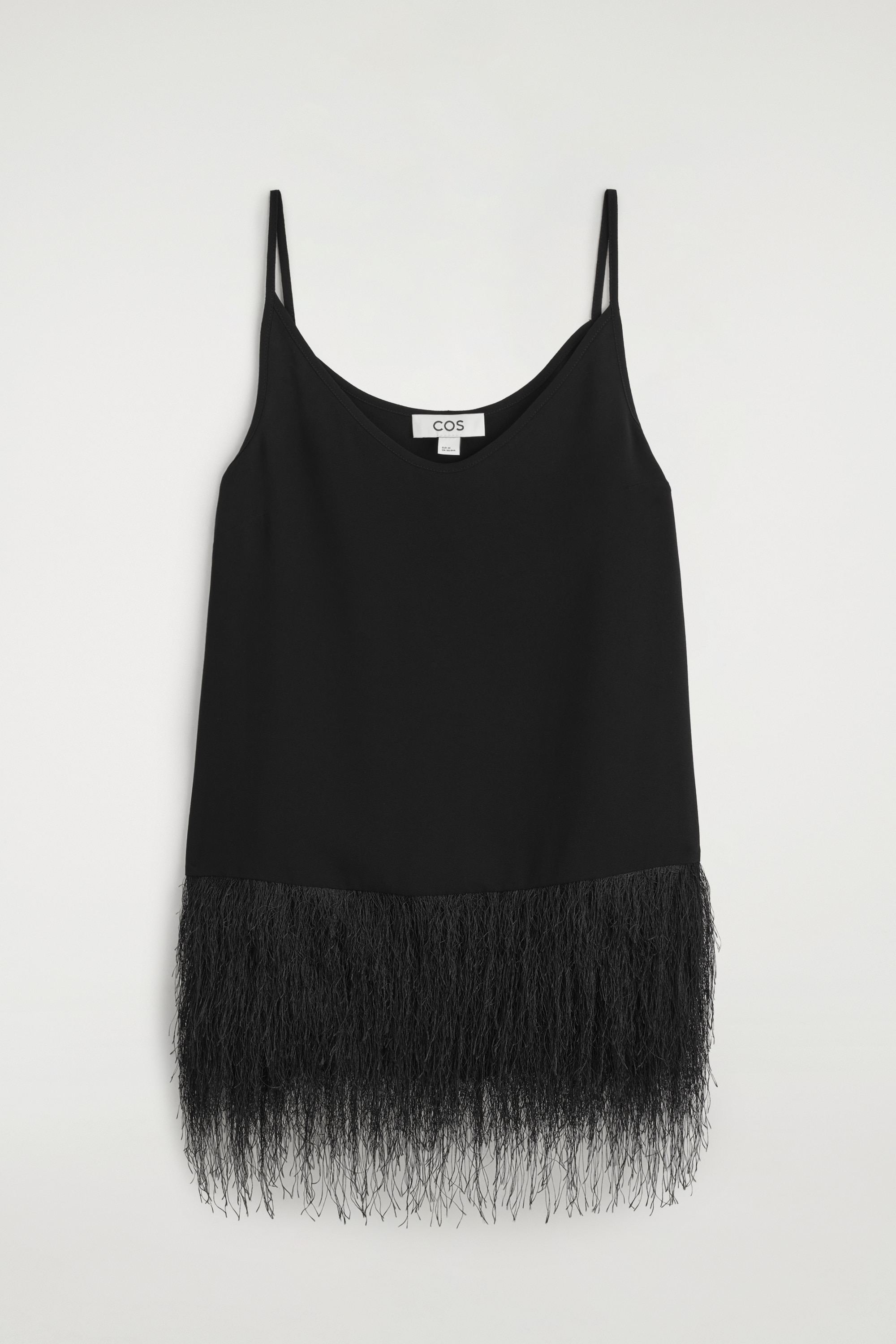A black cami top featuring a deep V-neckline, thin shoulder straps and a wide, textured fringe hem is worn by the model. The top is styled with black trousers, and the model is holding a black leather clutch.