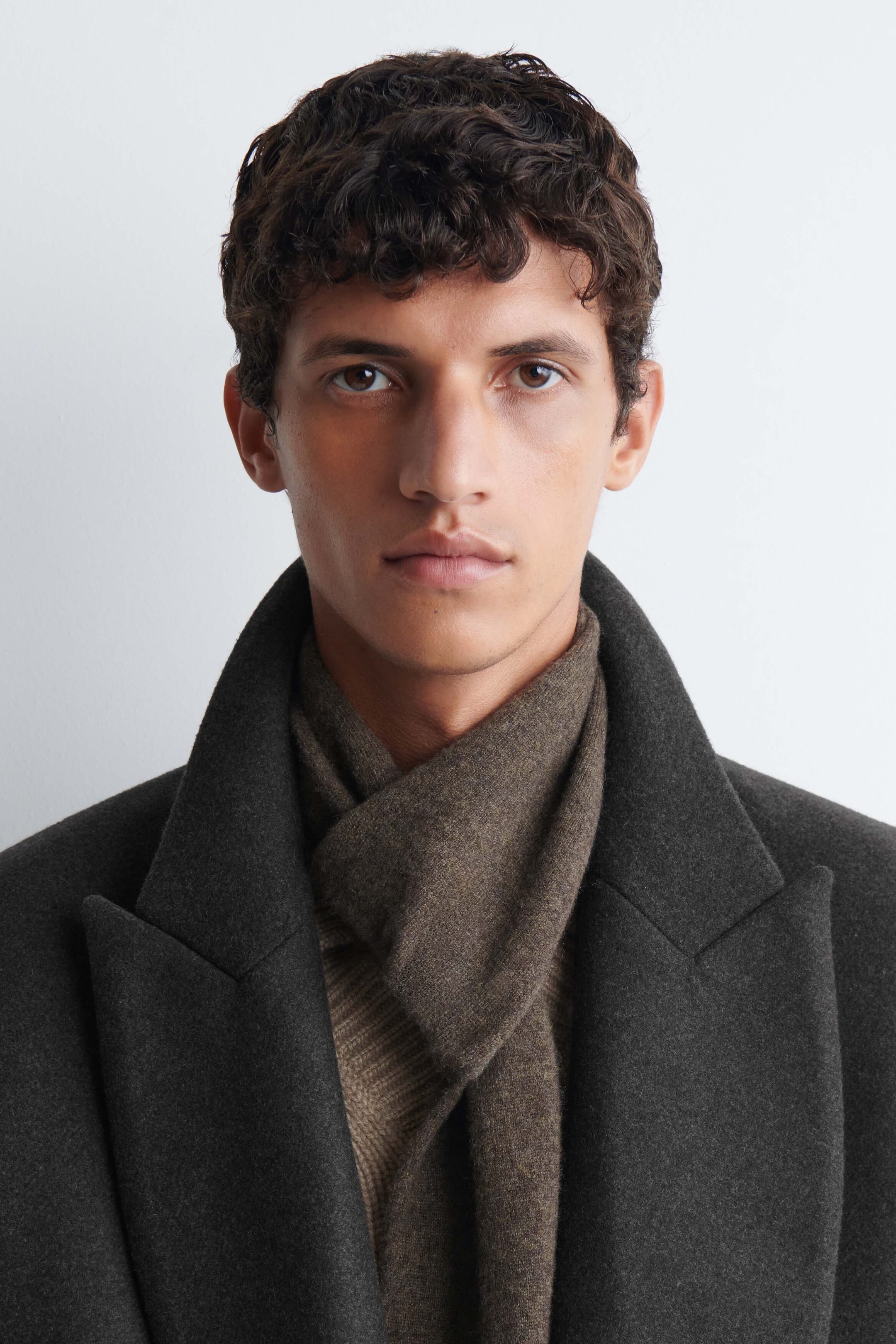 A brown cashmere pull-through scarf is worn wrapped around the model’s neck, with one end threaded through the loop at the front. The scarf is layered under a dark gray wool coat with wide lapels and over a brown ribbed knit top. A brown cashmere pull-through scarf is worn wrapped around the model’s neck, with one end threaded through the loop at the front. The scarf is layered under a dark gray wool coat with wide lapels and over a brown ribbed knit top.