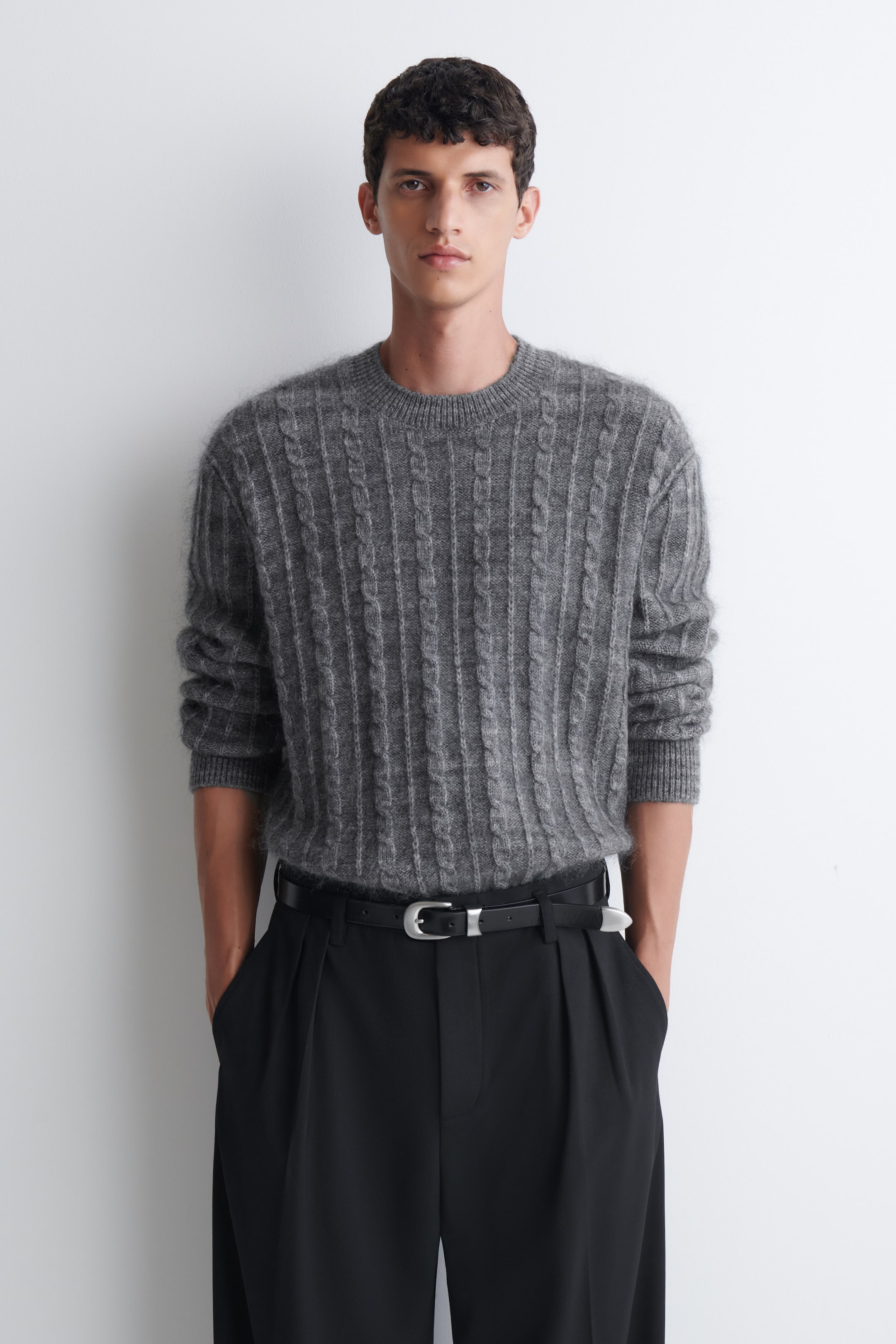 Visualizza l'immagine ingrandita: Un maglione grigio lavorato a trecce, con una finitura testurizzata e morbida, è indossato dalla modella. Il maglione presenta un girocollo e polsini a costine ed è infilato in pantaloni neri plissettati, fermati da una cintura nera.