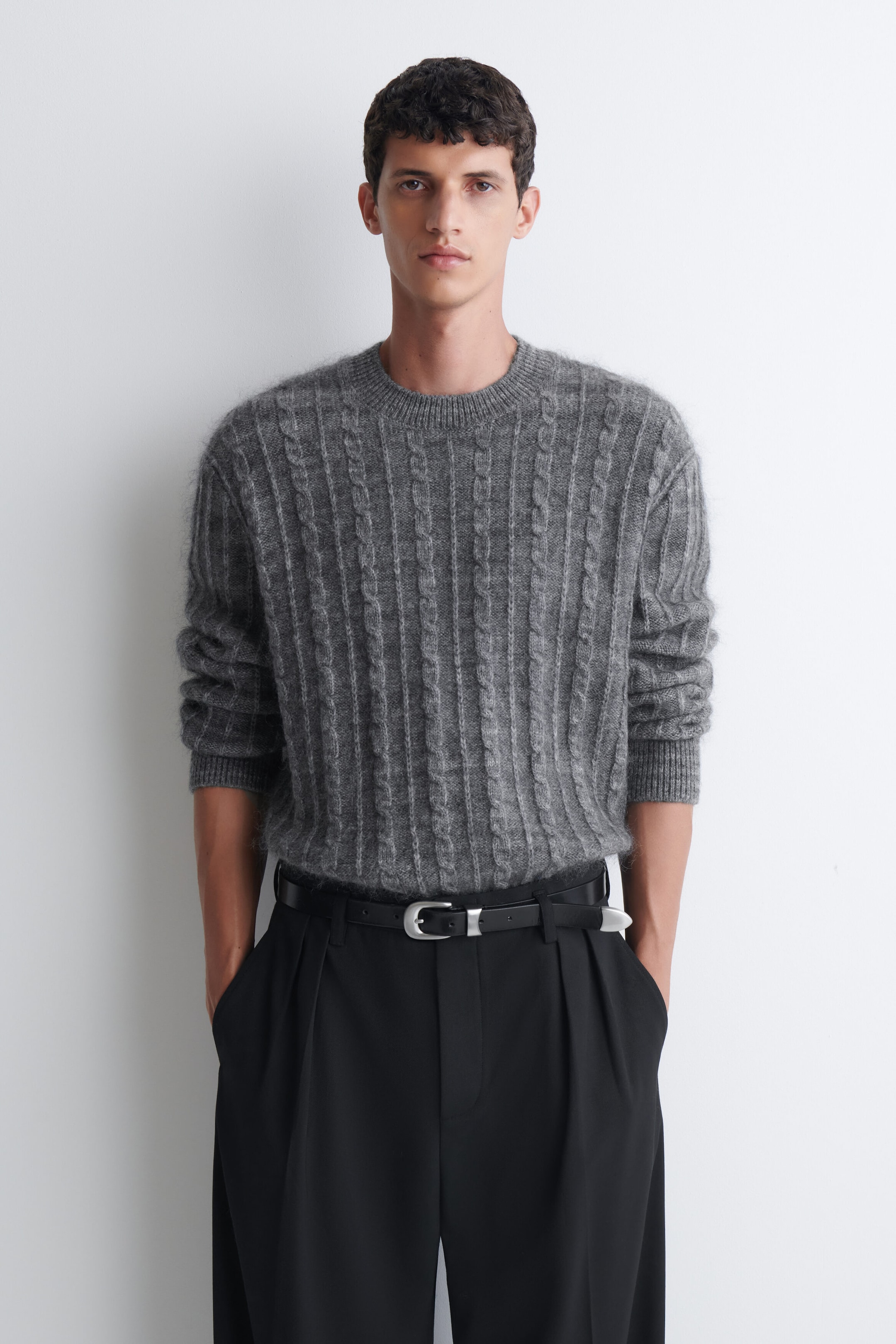 A grey cable-knit sweater with a textured, fuzzy finish is being worn by the model. The sweater features a ribbed crewneck and cuffs, and is tucked into black pleated trousers secured with a black belt. A grey cable-knit sweater with a textured, fuzzy finish is being worn by the model. The sweater features a ribbed crewneck and cuffs, and is tucked into black pleated trousers secured with a black belt.