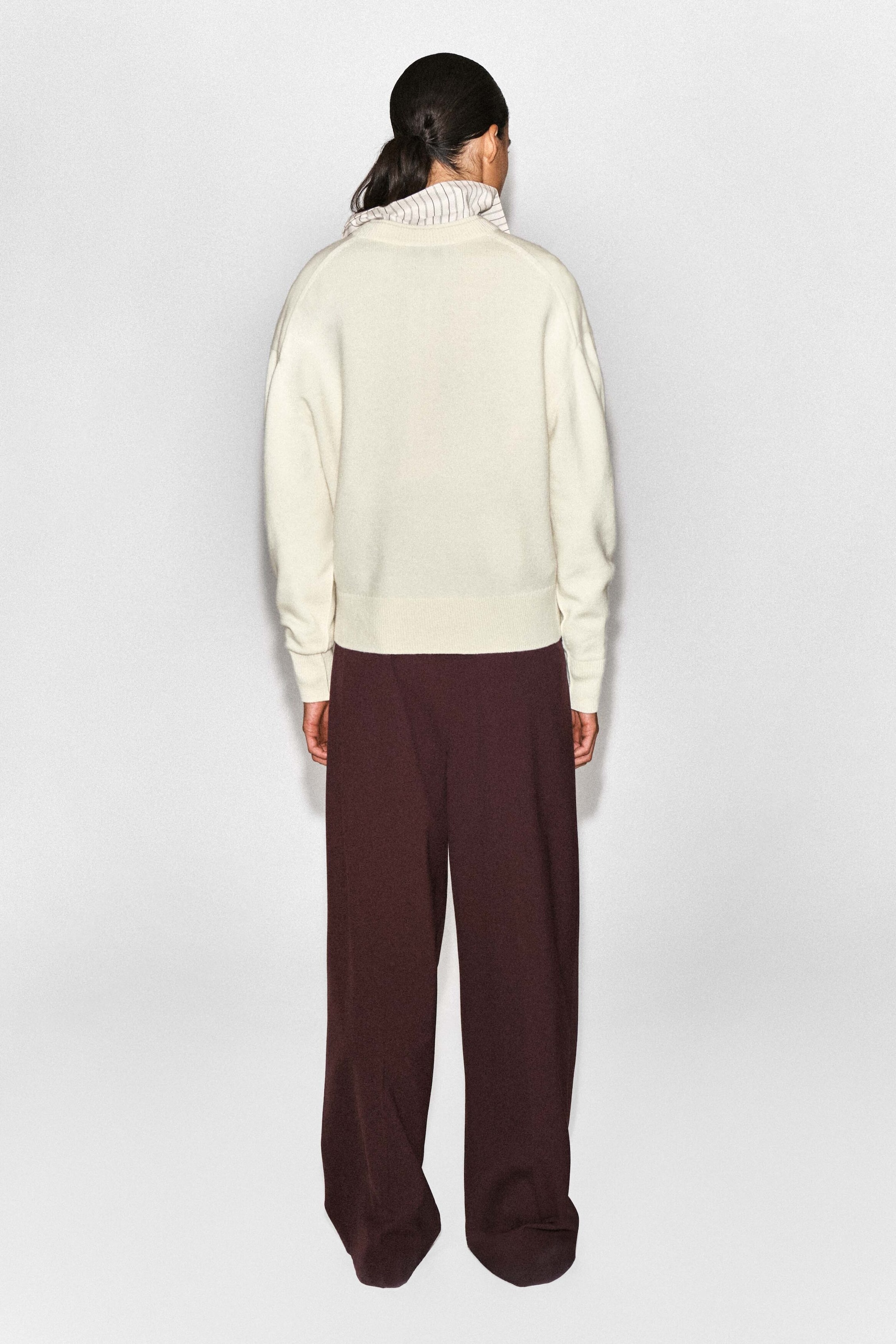 The cream-coloured merino wool layered V-neck jumper is shown from the back, highlighting its relaxed fit, ribbed hem, and ribbed cuffs. A striped shirt collar peeks out at the neckline, and the jumper is paired with dark burgundy wide-leg trousers. The cream-coloured merino wool layered V-neck jumper is shown from the back, highlighting its relaxed fit, ribbed hem, and ribbed cuffs. A striped shirt collar peeks out at the neckline, and the jumper is paired with dark burgundy wide-leg trousers.
