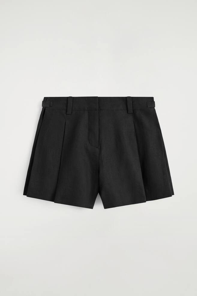 Schwarze Leinen-Shorts sind von vorne zu sehen und präsentieren eine weite Beinform, die durch Falten auf der Vorderseite verstärkt wird. Der Bund ist mit Gürtelschlaufen und verstellbaren Schnallenlaschen an jeder Seite ausgestattet und hat einen zentralen Reißverschluss. - 1