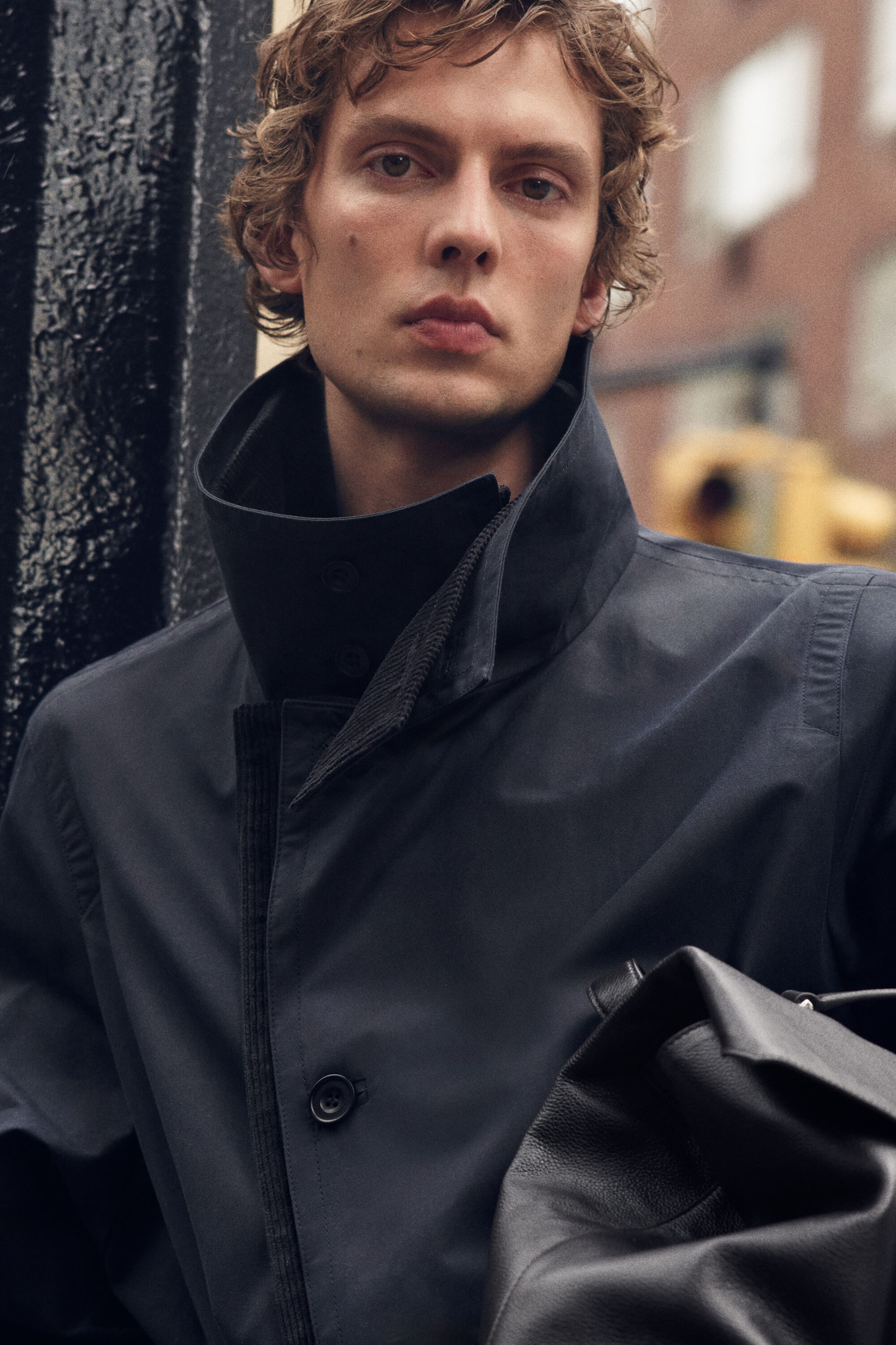 A close-up front view shows a dark navy waxed cotton trench coat featuring a high, structured funnel-neck collar and a button placket lined with tonal corduroy. The coat is worn with the collar turned up, while a textured black leather bag is held in the foreground. A close-up front view shows a dark navy waxed cotton trench coat featuring a high, structured funnel-neck collar and a button placket lined with tonal corduroy. The coat is worn with the collar turned up, while a textured black leather bag is held in the foreground.