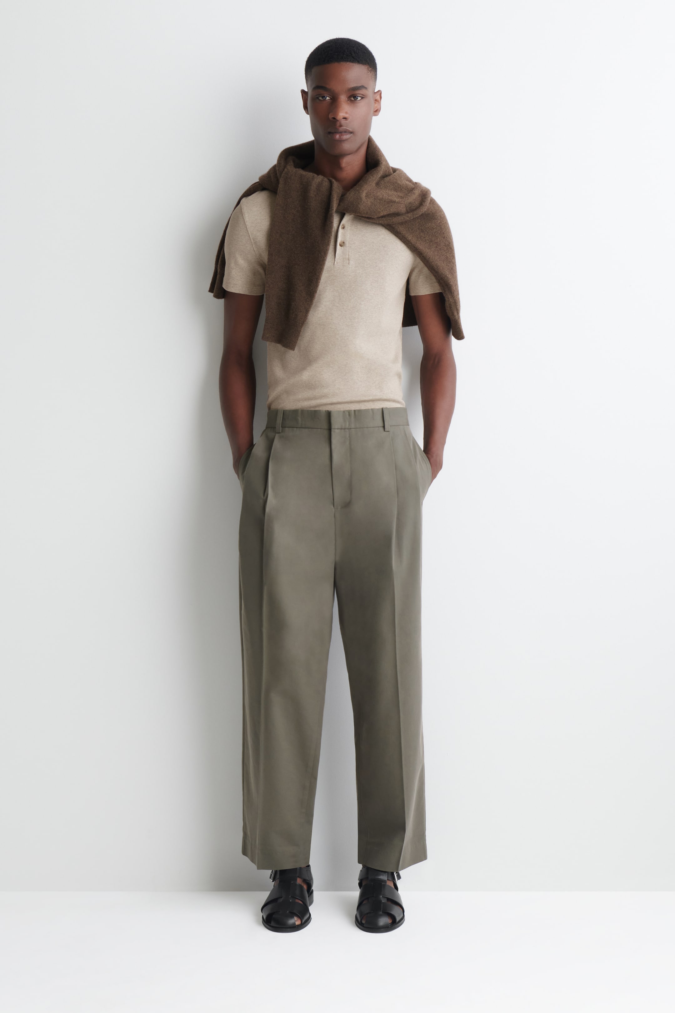 Vezi imaginea mai mare: Pantaloni conici de culoare verde măsliniu, cu croială relaxată, pliuri în față și găici pentru curea, purtați de model. Modelul își introduce mâinile în buzunarele laterale, iar pantalonii sunt asortați cu un polo băgat în pantaloni și sandale.