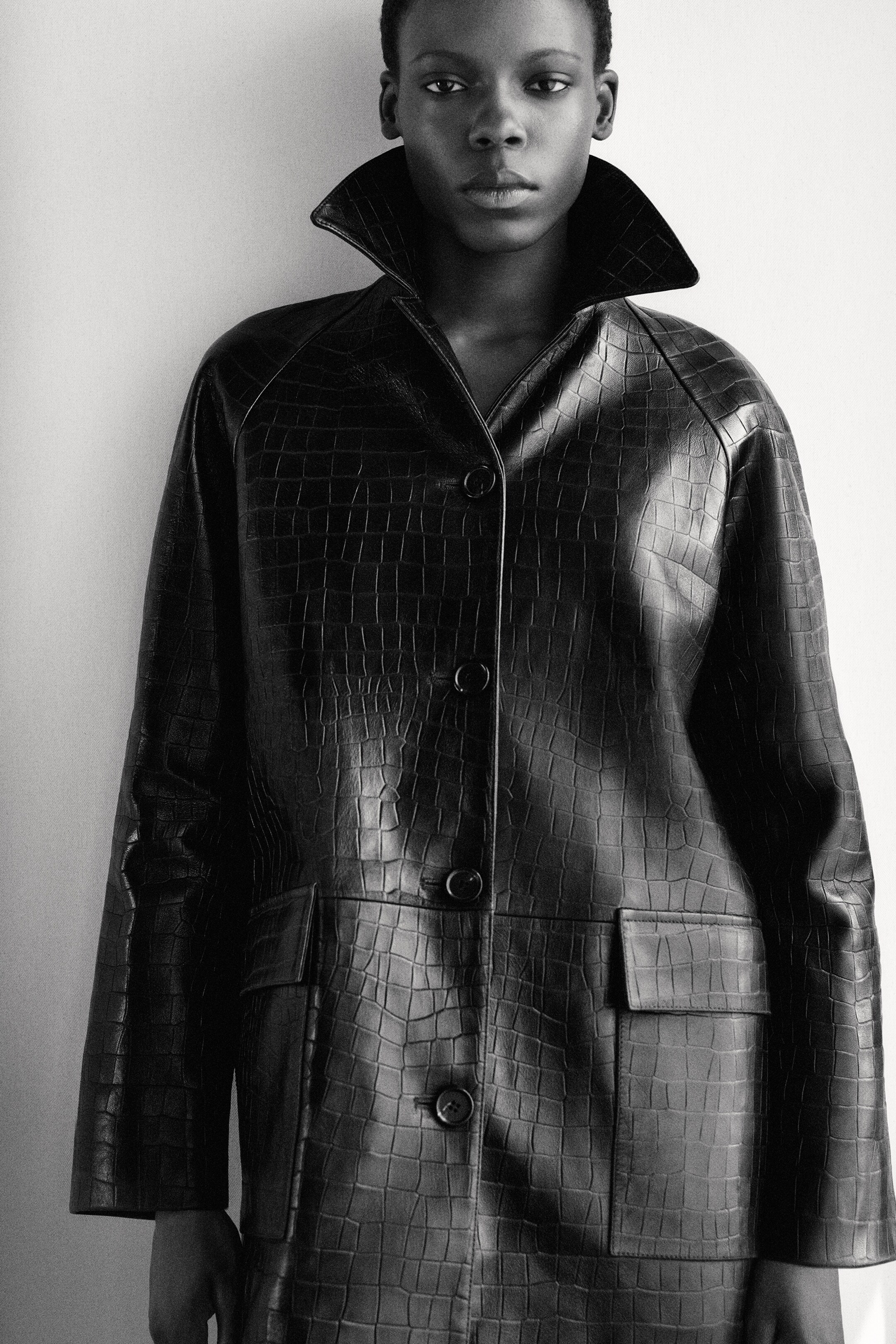 Größeres Bild anzeigen: Ein schwarzer Mantel mit Krokodilprägung verfügt über einen strukturierten, stehenden Spitzkragen, einen vorderen Knopfverschluss mit drei dunklen, runden Knöpfen und zwei großen, rechteckigen Klappentaschen an der unteren Vorderseite.