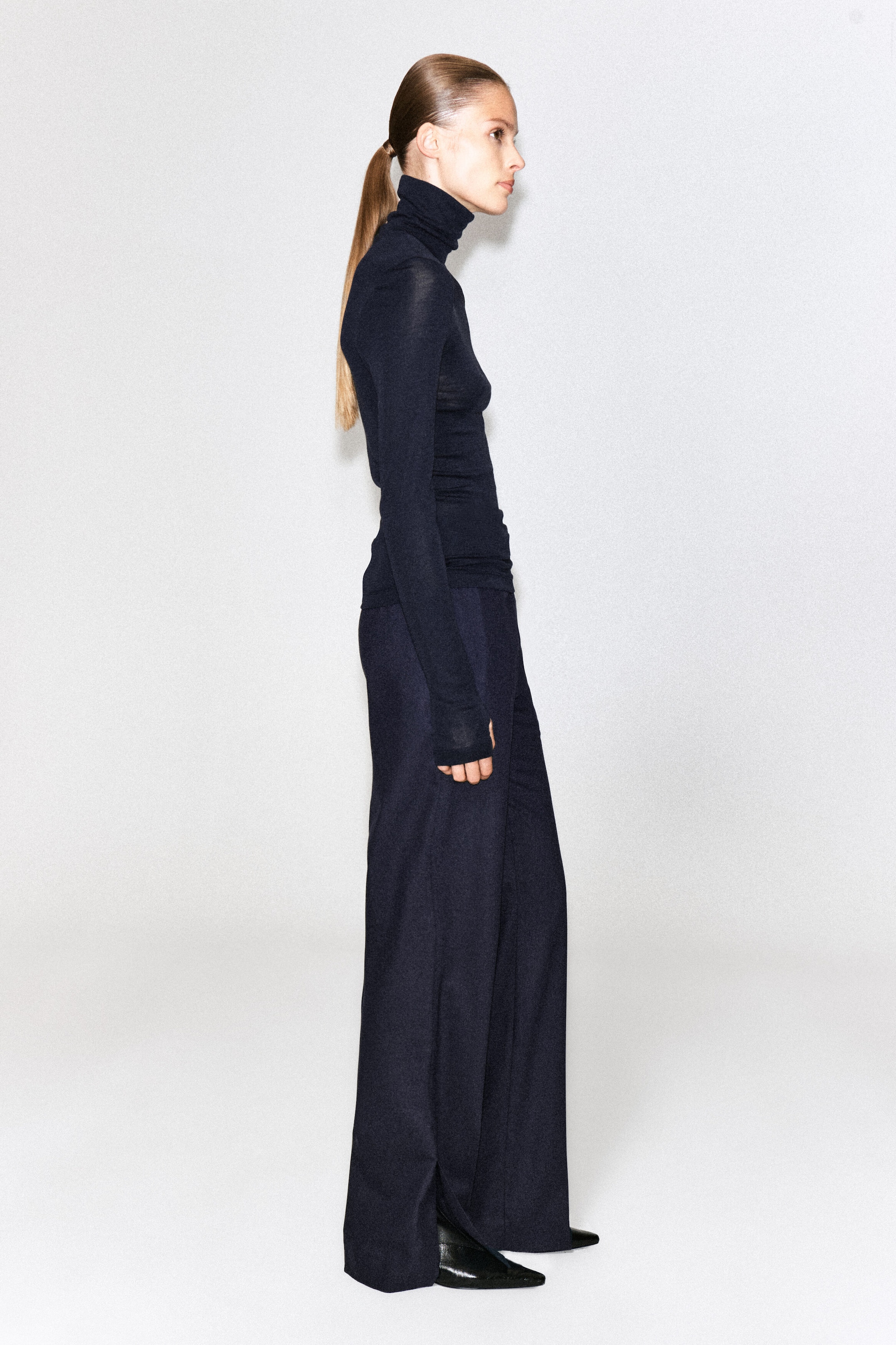 A dark navy merino wool high-neck top is being worn by the model, featuring a slim fit, long sleeves, and a folded high collar. The fine-knit top is styled with coordinating dark wide-leg trousers.