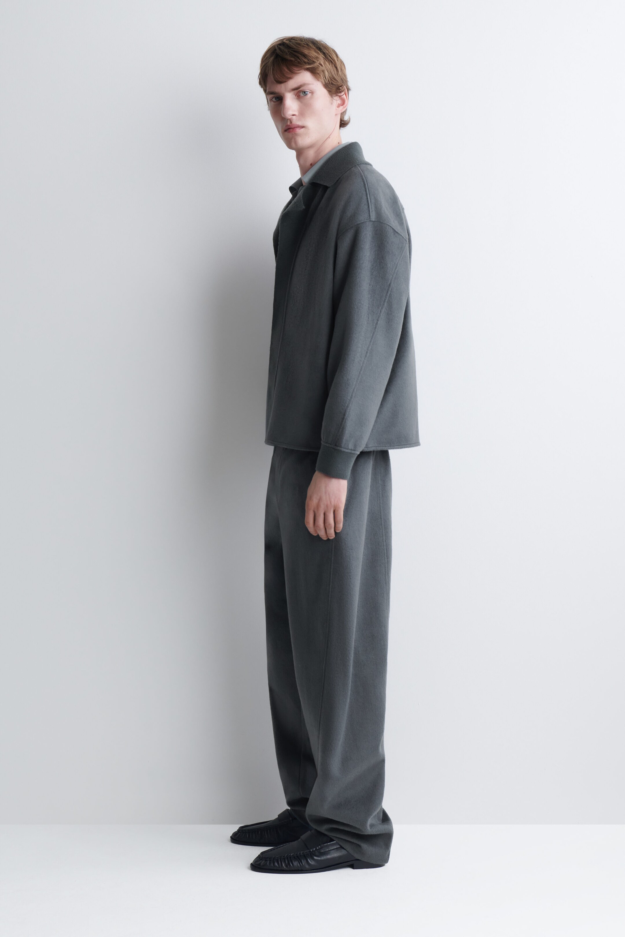 Le modèle porte un ensemble assorti composé d’une veste grise texturée et d’un pantalon à jambe large. La veste présente un col et des manches longues avec poignets côtelés, tandis que le pantalon à coupe ample tombe sur des chaussures noires en cuir. Le modèle porte un ensemble assorti composé d’une veste grise texturée et d’un pantalon à jambe large. La veste présente un col et des manches longues avec poignets côtelés, tandis que le pantalon à coupe ample tombe sur des chaussures noires en cuir.