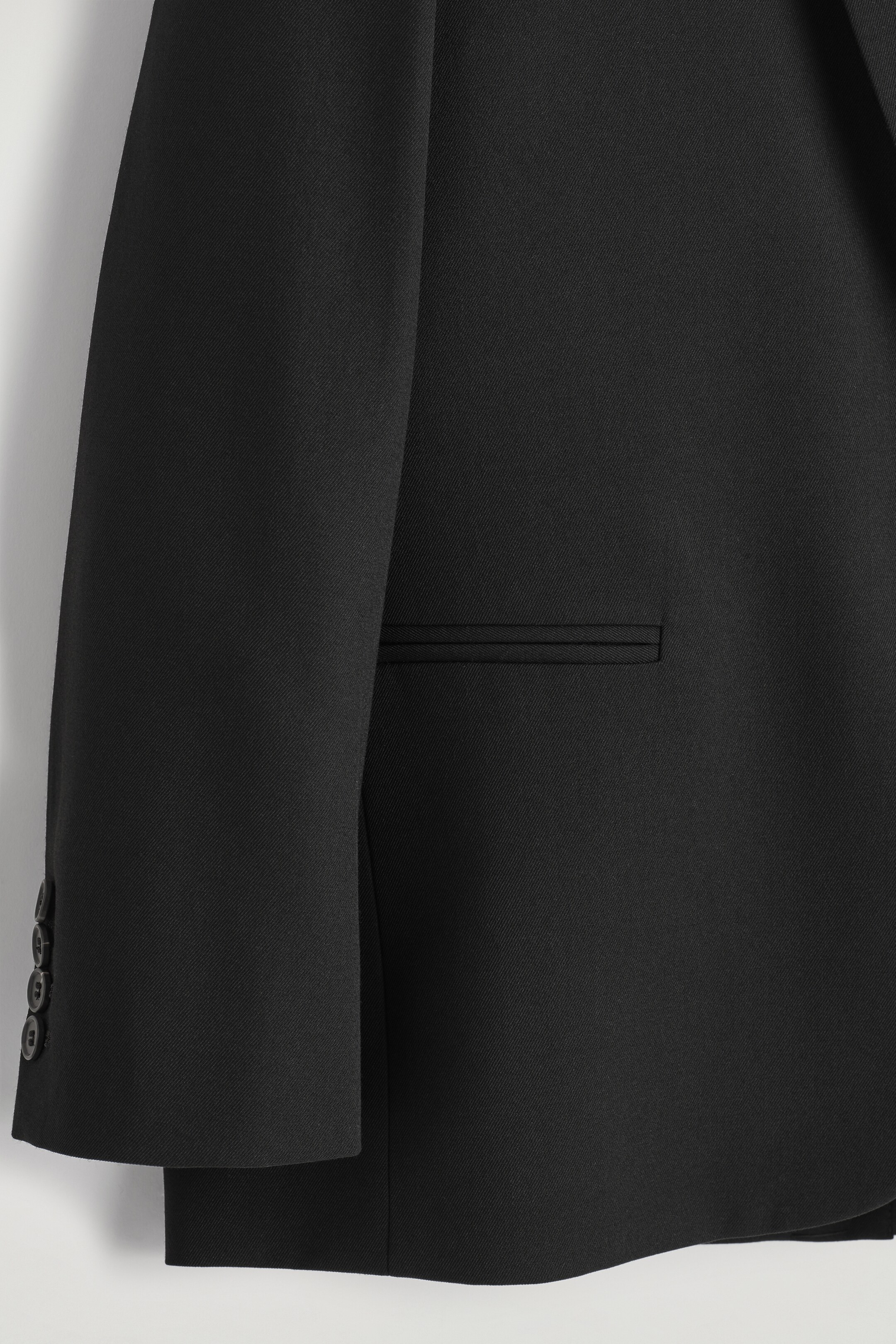 Größeres Bild anzeigen: Ein schwarzer Blazer ist in einer Nahaufnahme zu sehen, mit einer horizontalen Leistentasche auf dem Seitenpanel und drei dunklen, runden Knöpfen am Ärmelbündchen.