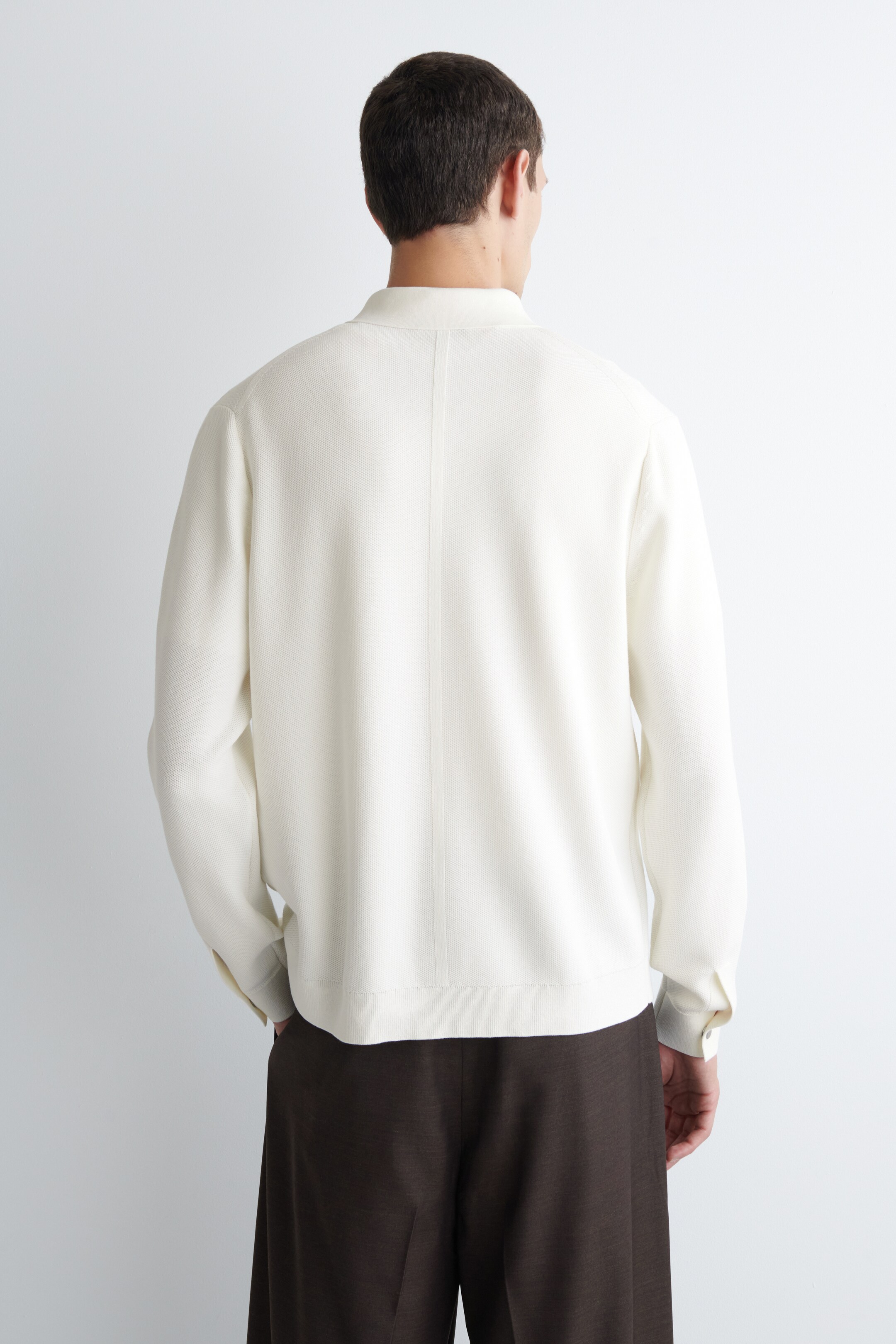 View larger image: A cream-colored knit shirt is being worn, viewed from the back. It is featuring long sleeves with buttoned cuffs and a stand collar. A vertical seam is running down the center back, and the shirt is falling over dark pants.