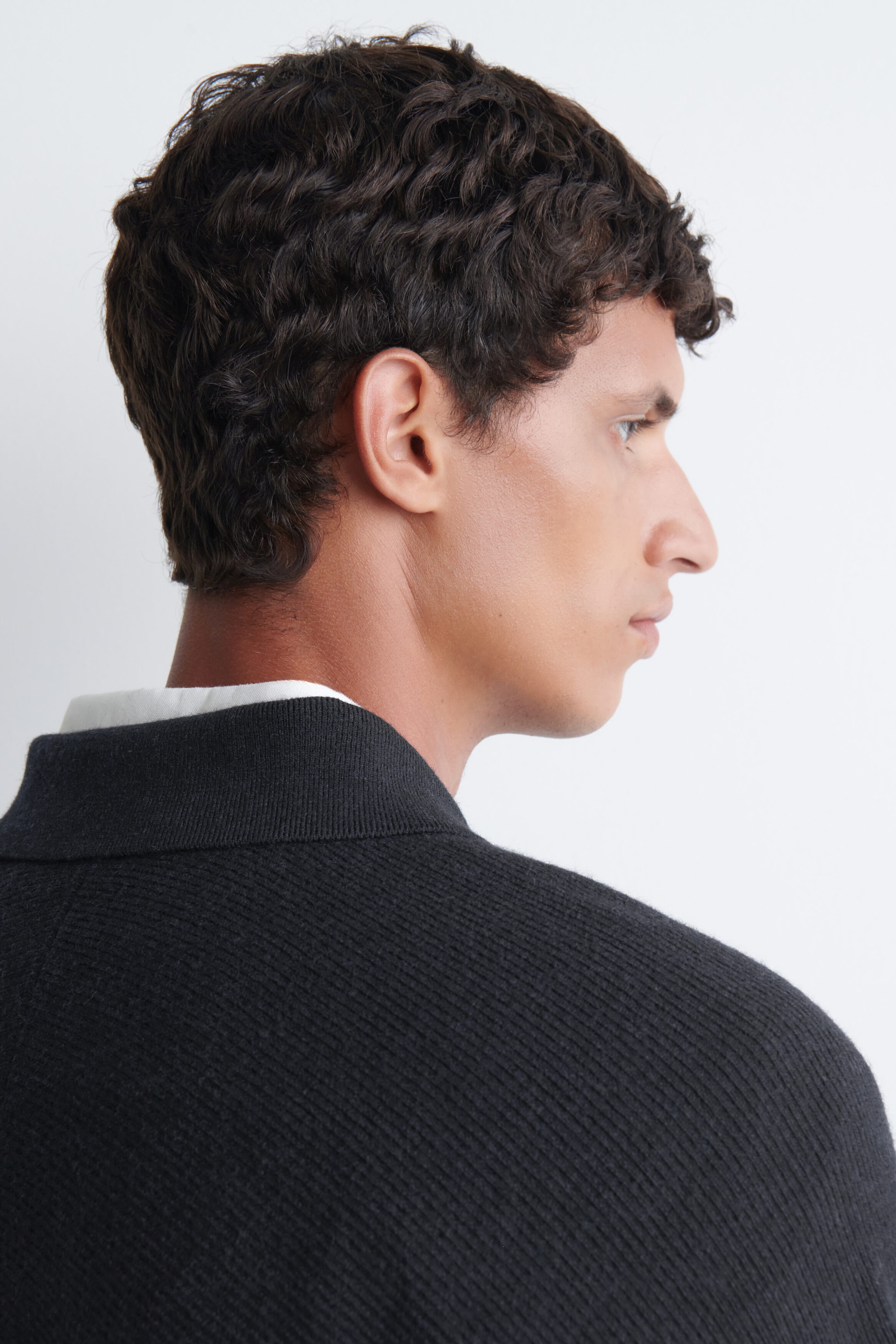 A dark grey boiled-wool jumper featuring a fine ribbed texture and a structured collar is being worn by the model, shown from a side-back profile. A white shirt collar is visible layered underneath the jumper's neckline. A dark grey boiled-wool jumper featuring a fine ribbed texture and a structured collar is being worn by the model, shown from a side-back profile. A white shirt collar is visible layered underneath the jumper's neckline.