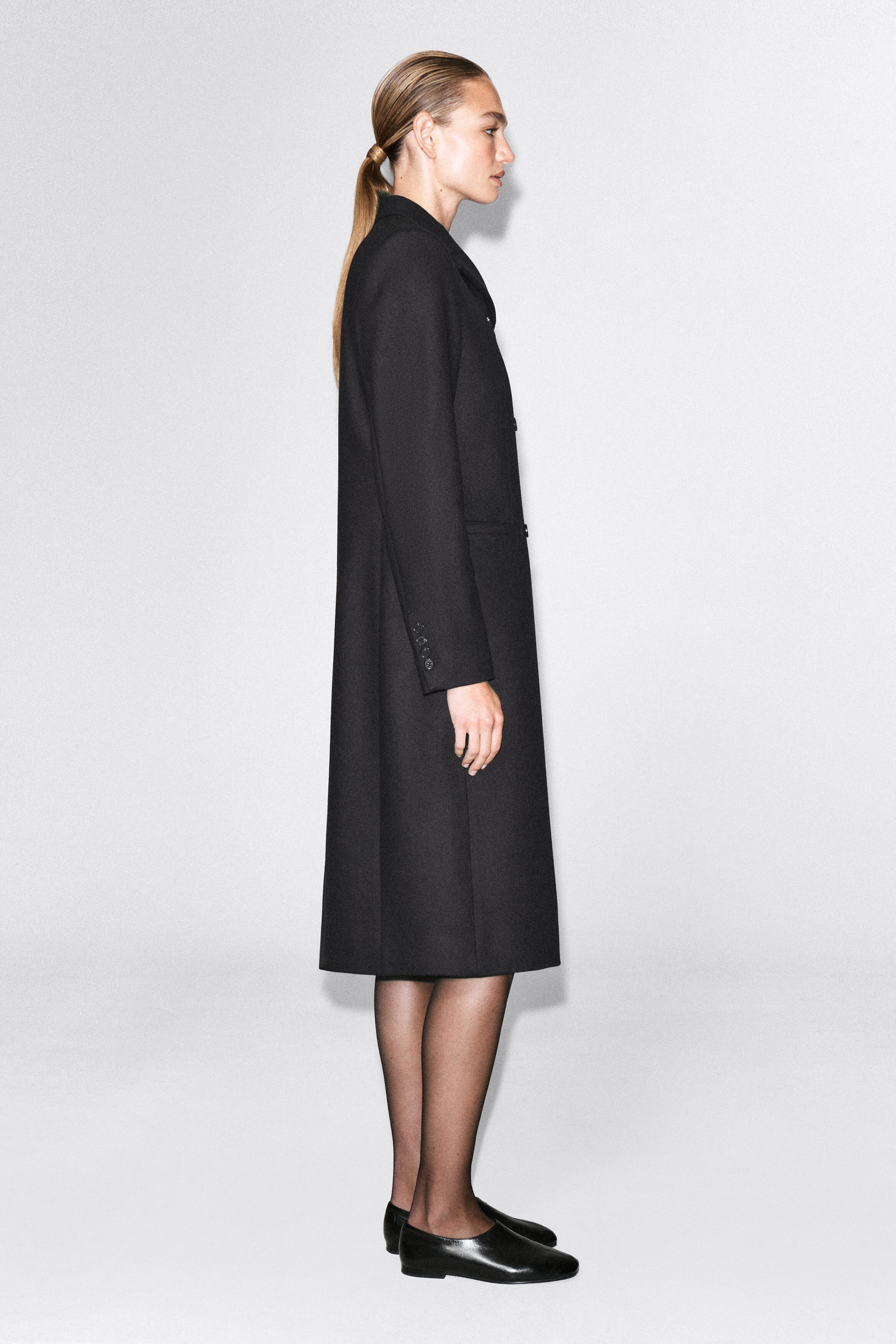 A slim-fit, longline black coat is being worn by the model, who is standing in a side-profile pose. The structured coat features long sleeves with buttoned cuffs and a midi length, styled with sheer black tights and black flat slip-on shoes. A slim-fit, longline black coat is being worn by the model, who is standing in a side-profile pose. The structured coat features long sleeves with buttoned cuffs and a midi length, styled with sheer black tights and black flat slip-on shoes.