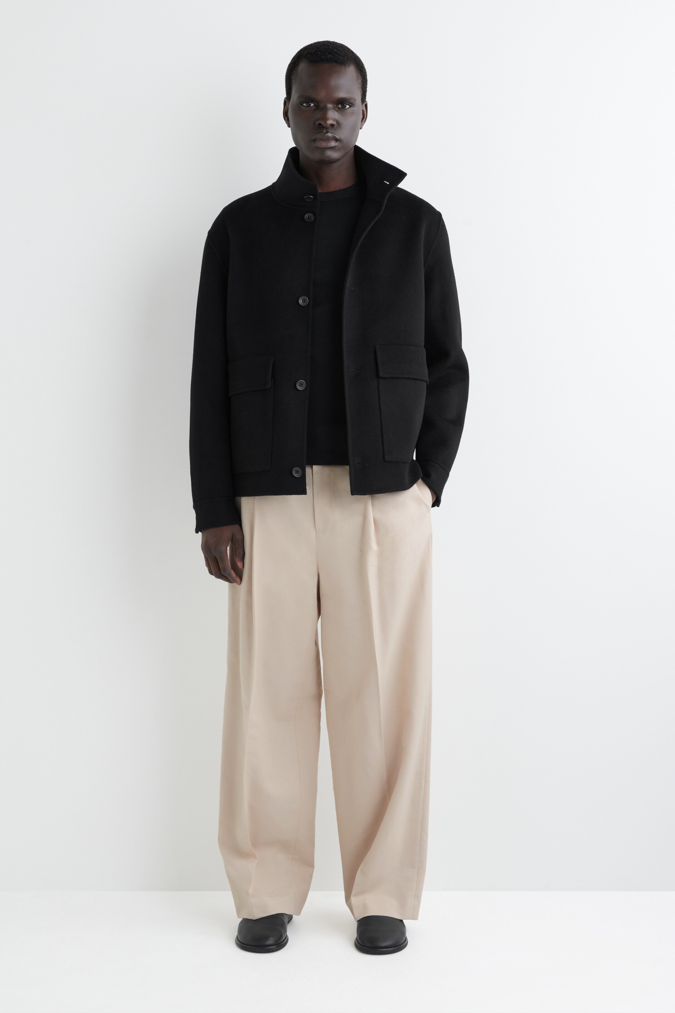 Ver imagen más grande: Se presenta una chaqueta negra, con un cuello alto, un cierre de botones en la parte delantera y dos grandes bolsillos de parche con solapa. Está superpuesta sobre una camiseta negra de cuello redondo y se combina con pantalones beige sueltos y plisados.