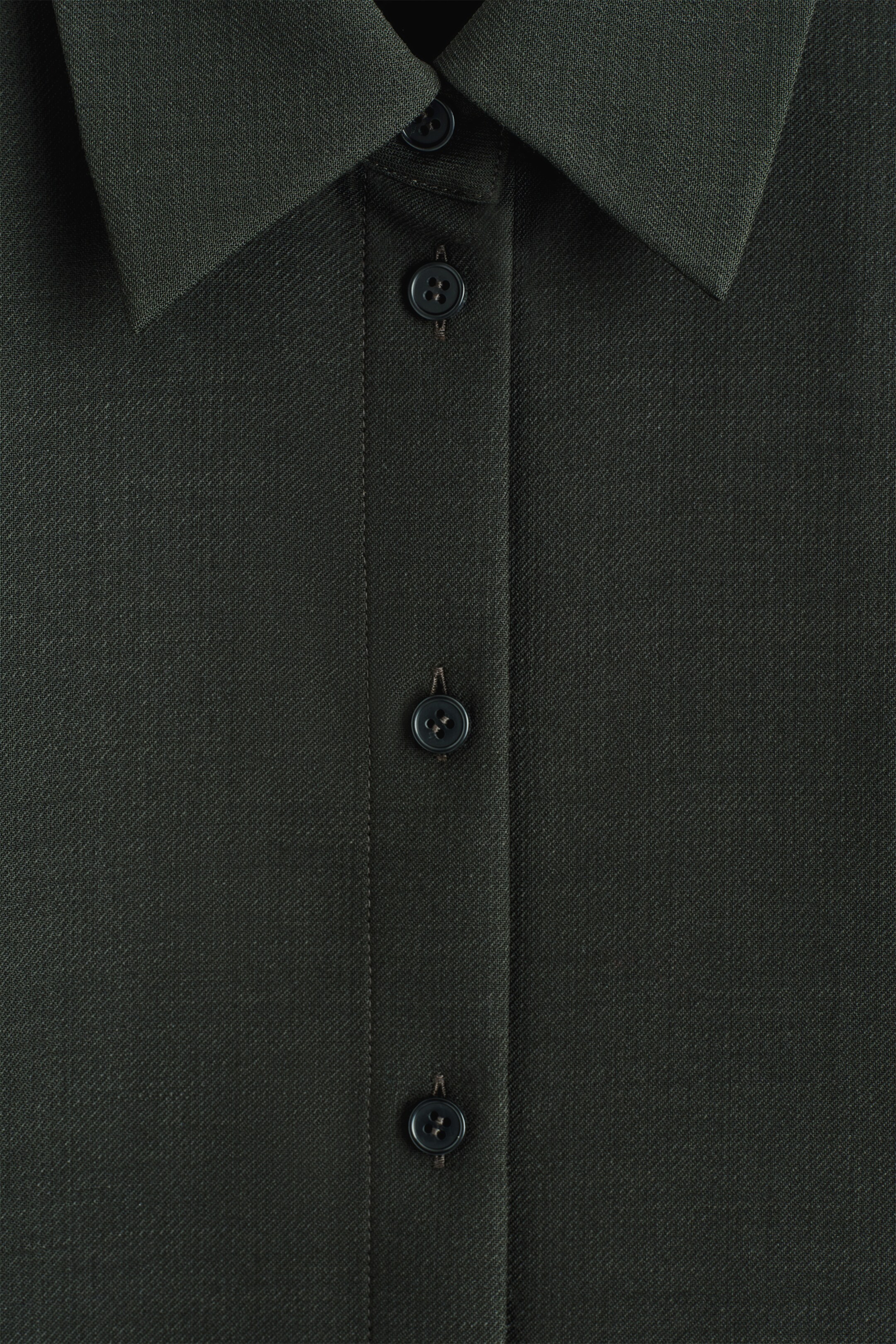 Grotere afbeelding bekijken: RELAXED WOOL SHIRT. Het donkergroene wollen hemd wordt van voren gepresenteerd, waarbij de subtiele keperweefseltextuur prominent wordt getoond. Een klassieke puntkraag omlijst de halslijn, en een centrale knopenlijst loopt over de voorkant, met drie donkere, ronde knopen die het vastmaken.