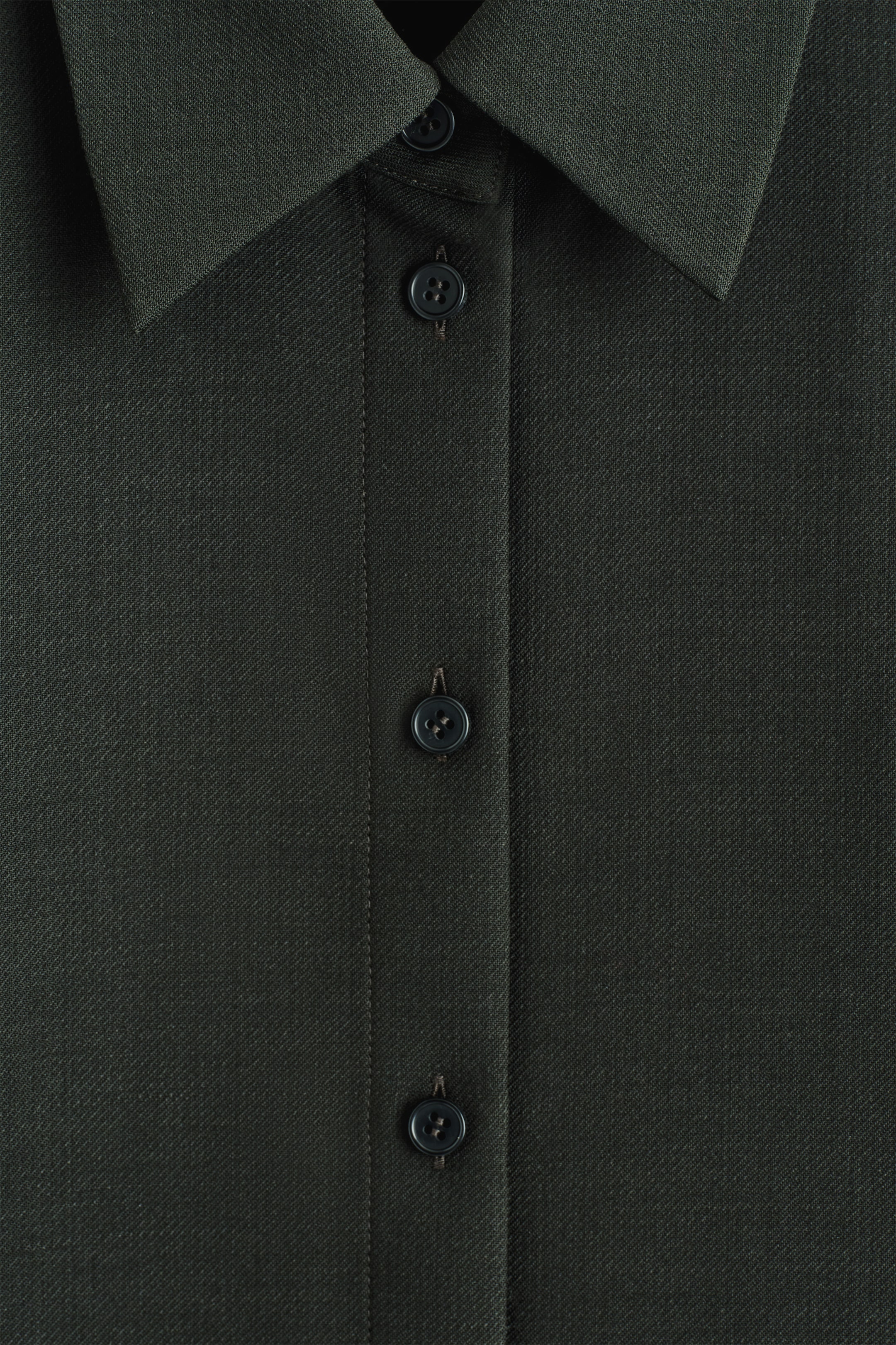 Agrandir l'image: CHEMISE EN LAINE DÉCONTRACTÉE. La chemise en laine vert foncé est présentée de face, mettant en avant sa texture subtile de tissage sergé. Un col à pointes classique encadre l'encolure, et une patte de boutonnage centrale descend le long du devant, avec trois boutons ronds et foncés pour la fermer.