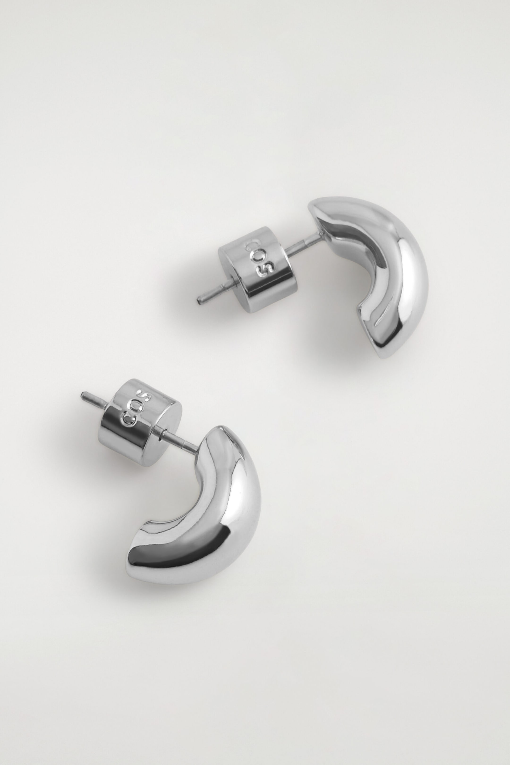 Des boucles d’oreilles demi-créoles sculpturales mettent en valeur un design en C courbé et volumineux, avec une finition argentée polie et très brillante. La paire est posée à plat, montrant des tiges cylindriques épaisses avec fermoirs à l’arrière, gravées du nom de la marque « COS ». Des boucles d’oreilles demi-créoles sculpturales mettent en valeur un design en C courbé et volumineux, avec une finition argentée polie et très brillante. La paire est posée à plat, montrant des tiges cylindriques épaisses avec fermoirs à l’arrière, gravées du nom de la marque « COS ».
