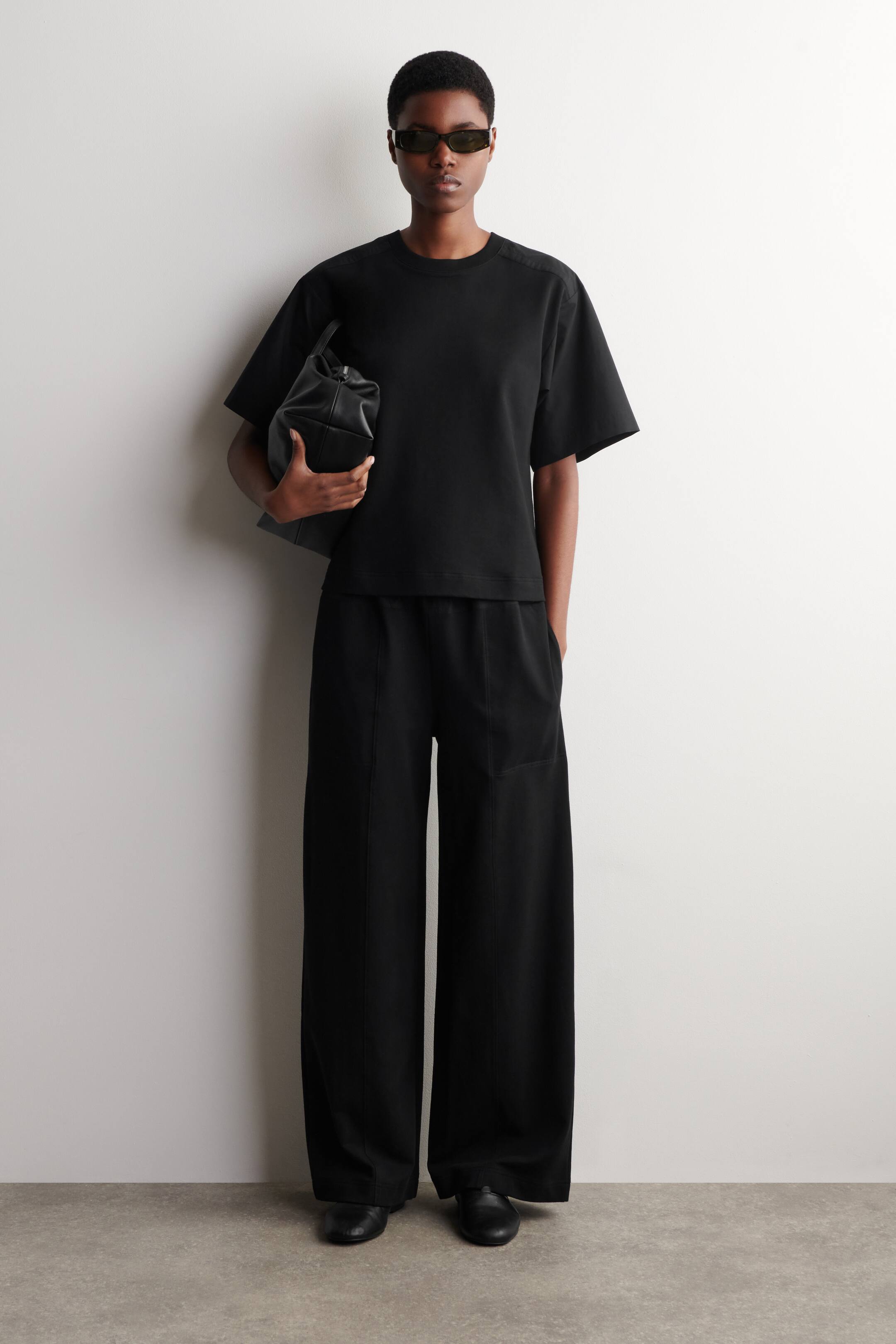 Größeres Bild ansehen: Ein schwarzes T-Shirt und eine Hose mit weitem Bein werden präsentiert. Das T-Shirt zeichnet sich durch einen Rundhalsausschnitt, kurze weite Ärmel und überschnittene Schulternähte aus, während die dazu passende Hose einen elastischen Bund und vertikale Frontnähte aufweist, die eine Silhouette mit weitem Bein formen.